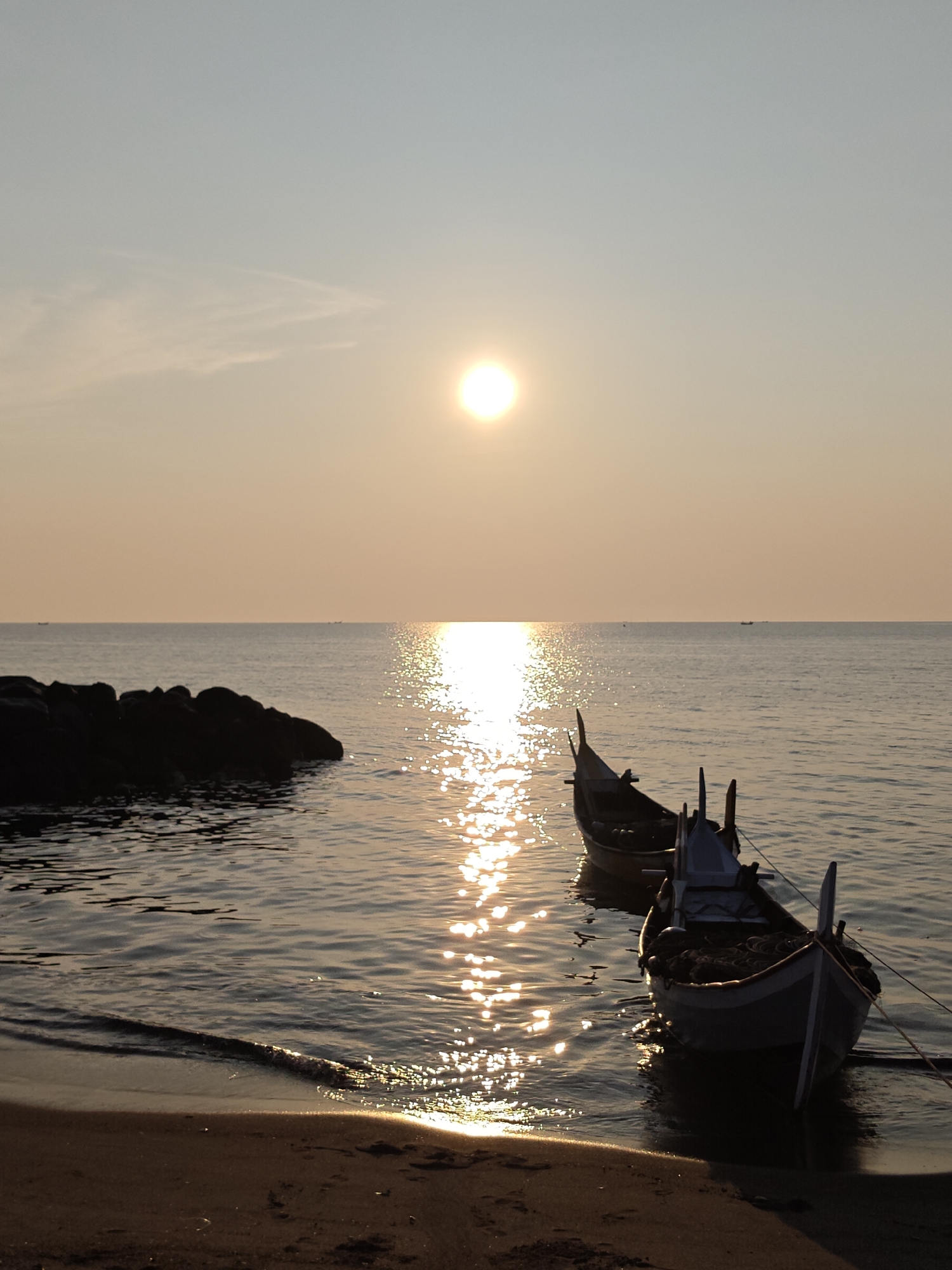 A serene coastal landscape captures a sunrise or sunset over the ocean in Lhokseumawe, Indonesia. The sun, a bright, luminous disc, sits low in the sky, casting a warm golden glow across the scene. Its intense light creates a shimmering path of reflections stretching across the calm, dark blue-grey water, extending towards the viewer.
In the foreground, two traditional wooden fishing boats are gently moored near the sandy shore, partially on the beach and partially in the shallow water. Their dark silhouettes are highlighted by the sun's glare on the water. The boat closer to the viewer appears to contain fishing nets. On the left side of the frame, a cluster of dark rocks juts out into the water, providing a contrasting element to the smooth horizon.
The sky above is a gradient of soft hues, transitioning from a pale blue-grey at the top to a warm, hazy orange-yellow near the horizon, with faint, wispy clouds barely visible. The water is largely still, with gentle ripples near the shoreline where small waves lap at the dark, wet sand. A faint, distant landmass or other boats can be seen on the horizon. The overall atmosphere is peaceful and tranquil, devoid of any visible human presence or text.