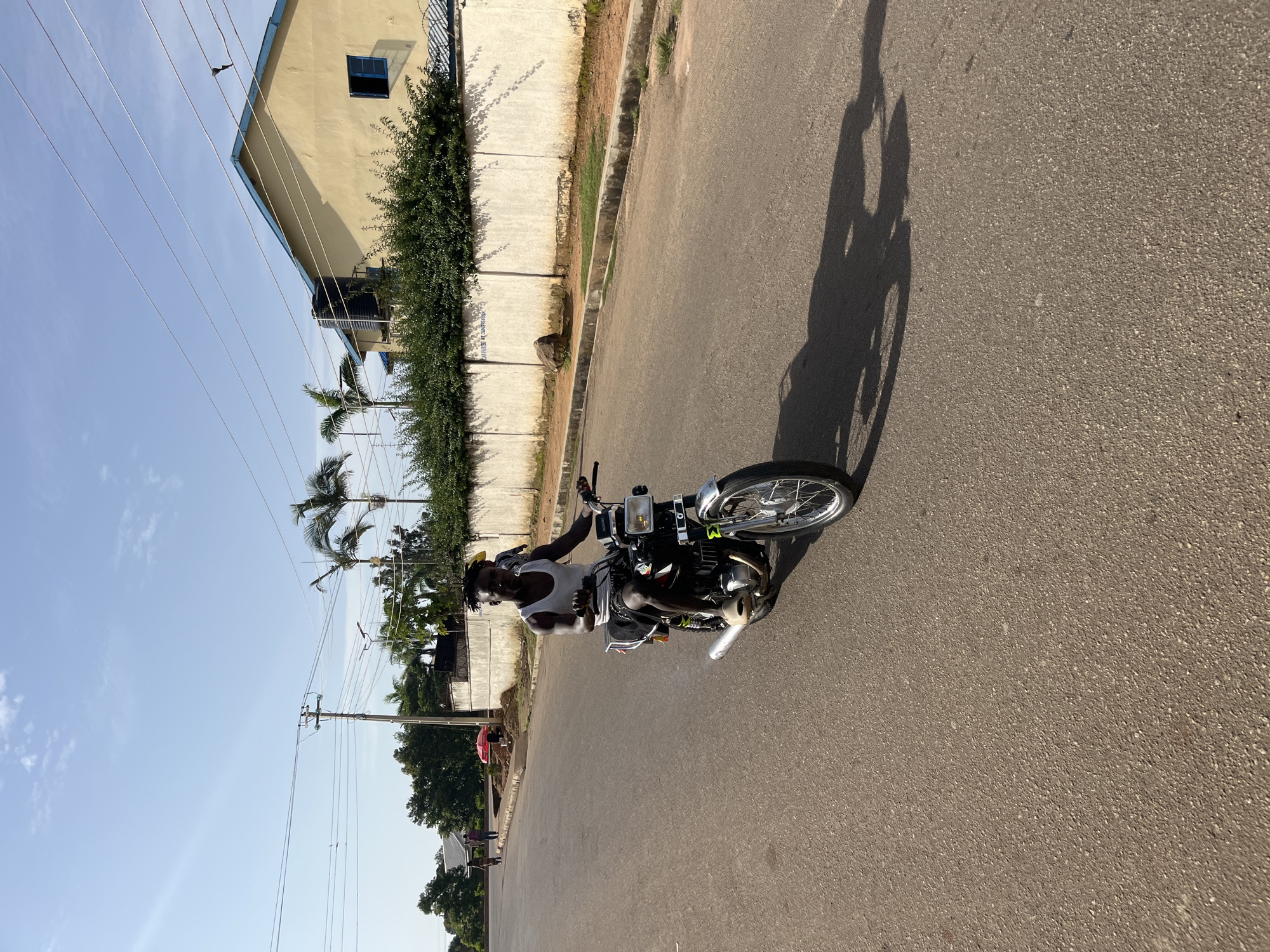 In Bukuru, Nigeria, a young Black man with dreadlocks rides a black Suzuki motorcycle. He is wearing a white tank top and dark shorts, and appears to be moving forward on a paved road. The man is in focus, and the background shows a wall, buildings, palm trees, and power lines under a clear blue sky with some clouds. The sun is casting a long shadow of the motorcycle and rider on the road. There are a few other people and a red umbrella visible in the distance to the left, suggesting a mildly populated area.