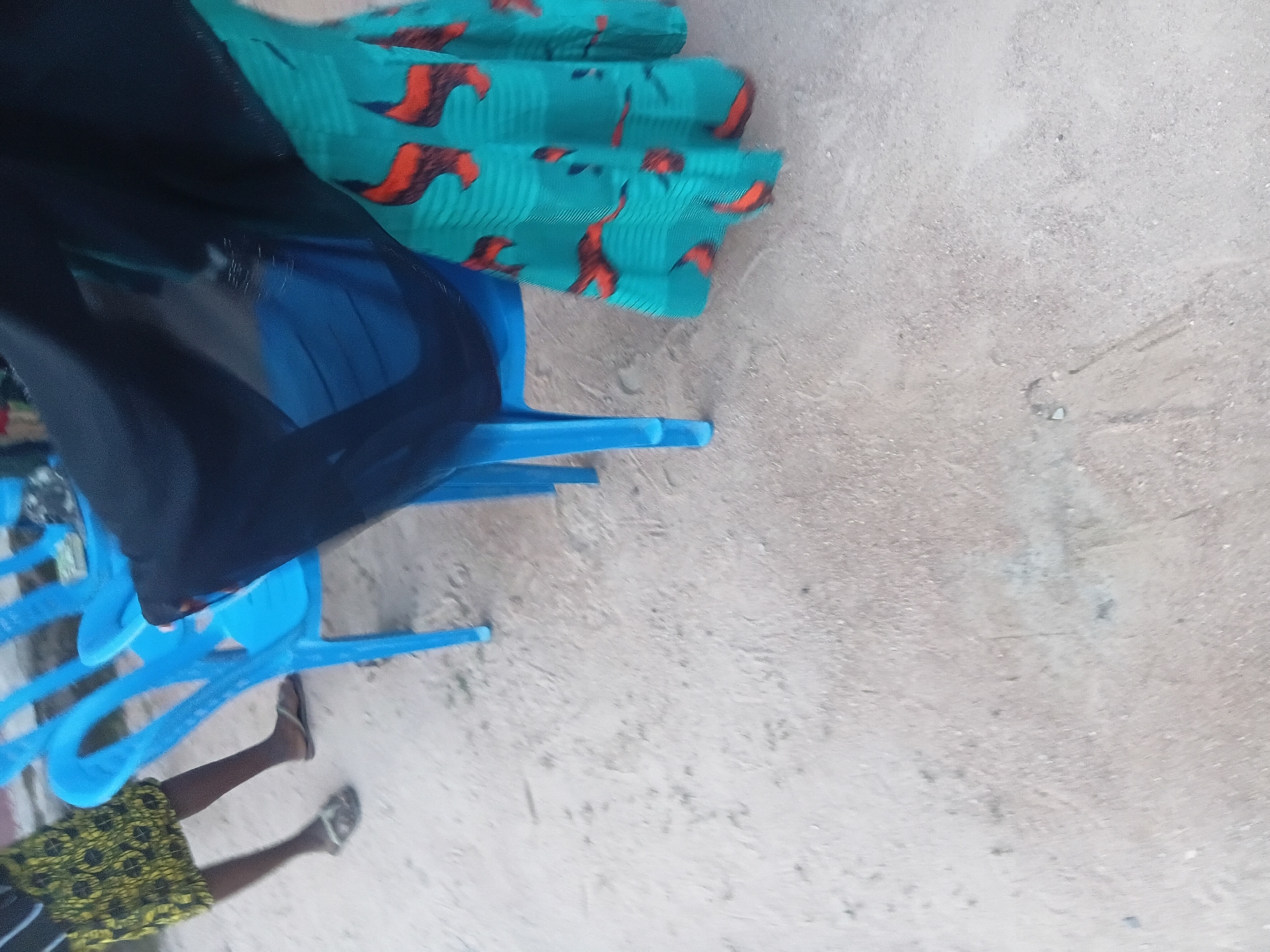A low-angle, medium shot captures two individuals partially visible in a sandy, outdoor environment. On the left, a person wearing a yellow and black patterned skirt and dark sandals stands with their legs bent. To the right, another individual is seated on a bright blue plastic chair. This person is covered by a flowing black garment, obscuring most of their body, but their lower half is clad in a vibrant turquoise skirt adorned with a red and black pattern resembling stylized flames or animal figures. The skirt is pleated and drapes around the legs and the blue chair. The ground is covered in light-colored sand with scattered small pebbles and debris. In the background, parts of other blue plastic chairs are visible, suggesting an outdoor seating area. The overall lighting is somewhat dim, indicating it might be overcast or late in the day. The scene is set in Jalingo, Nigeria.
