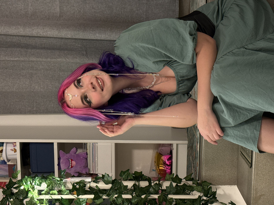 A woman with pink and purple hair wears a green dress and stands next to a white bookshelf adorned with artificial ivy. She wears a jeweled headpiece and matching necklace. The bookshelf holds items including a purple plush toy, a blue box, and balloons. A gray curtain is visible in the background. The image is rotated counterclockwise.