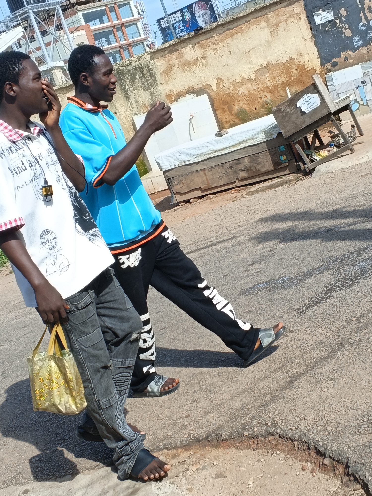 Two young men walk on a paved road in Jos, Nigeria. The man on the left wears a white t-shirt with graphics and a red and white checkered collar, grey cargo pants, and black sandals. He carries a small, yellow, sequined bag in his right hand. The man on the right wears a bright blue t-shirt with orange trim and black athletic pants with white lettering. He wears grey and black slide sandals. The background shows a dilapidated building with peeling paint and posters on its exterior wall, and a construction site with a large wooden structure covered by a white tarp. The road surface is cracked and worn, with shadows cast by the men and the structures.