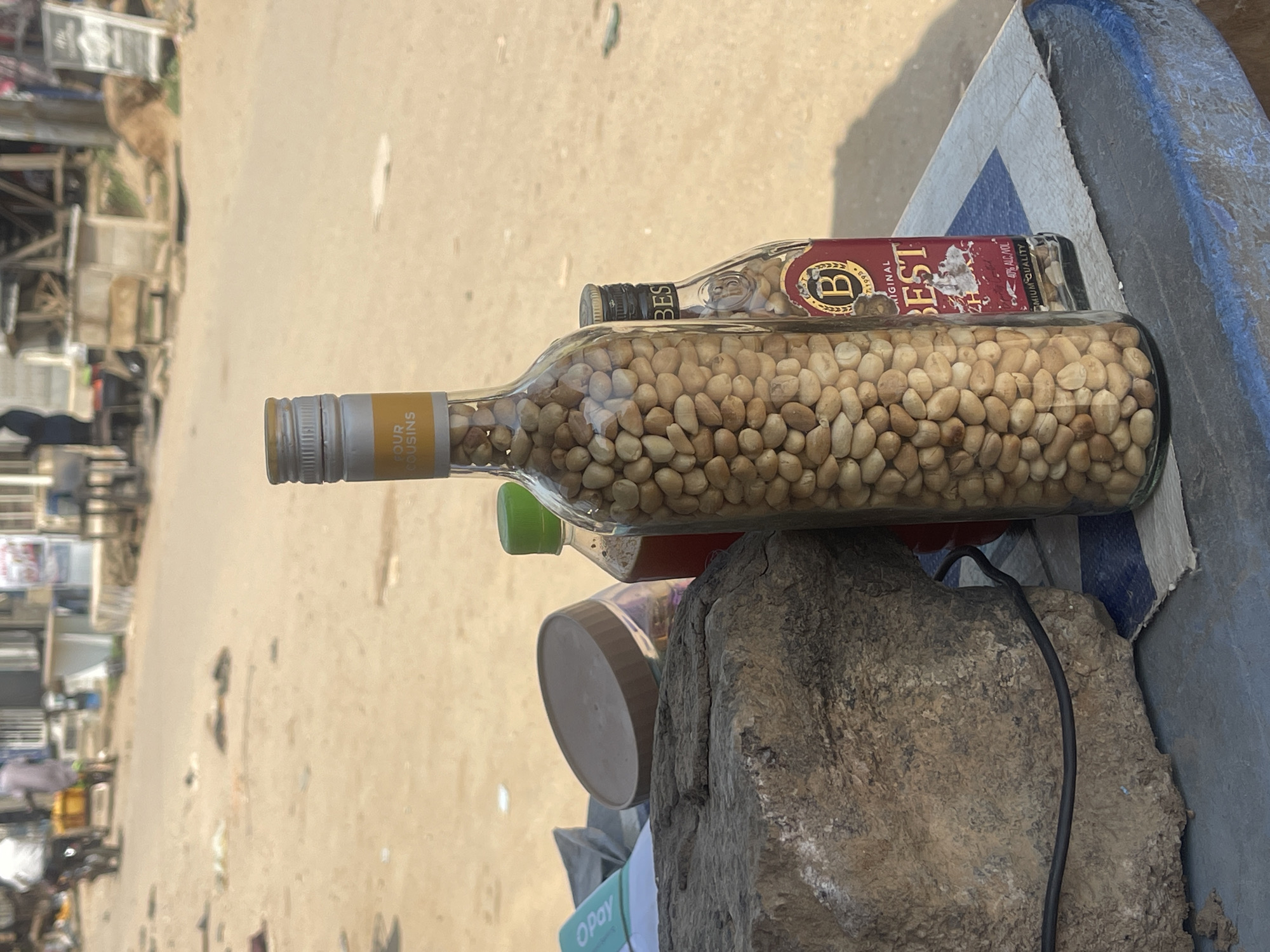 This media file shows an outdoor market scene in Abuja, Nigeria. The primary subjects are two glass bottles filled with small, light-brown nuts, possibly peanuts. The bottle in the foreground has a silver cap and a yellow label that reads "GOUR COUSINS". Behind it, a smaller bottle with a dark cap contains more nuts and has a red and gold label with the letter "B" and the text "BEST", "ORIGINAL", and "PREMIUM QUALITY". The bottles are placed on a rough, greyish-blue surface, possibly a table or stall, next to a large, rough rock.
In the background, a sandy or dusty ground is visible, with indistinct shapes that appear to be market stalls or structures. There are hints of people in the far background, suggesting a busy marketplace. The lighting indicates it is daytime, and the sand and open setting suggest warm, possibly dry weather. A plastic bottle with a green cap is also partially visible between the two main bottles. A crumpled green and white item with "OPay" text is lying near the rock. The scene appears to be a casual setup for selling goods in an open-air market.
Notable details include the specific text on the bottle labels, the texture of the rock, and the presence of the "OPay" branding, which is a mobile payment service popular in Nigeria. The overall impression is that of a vibrant, everyday market environment.