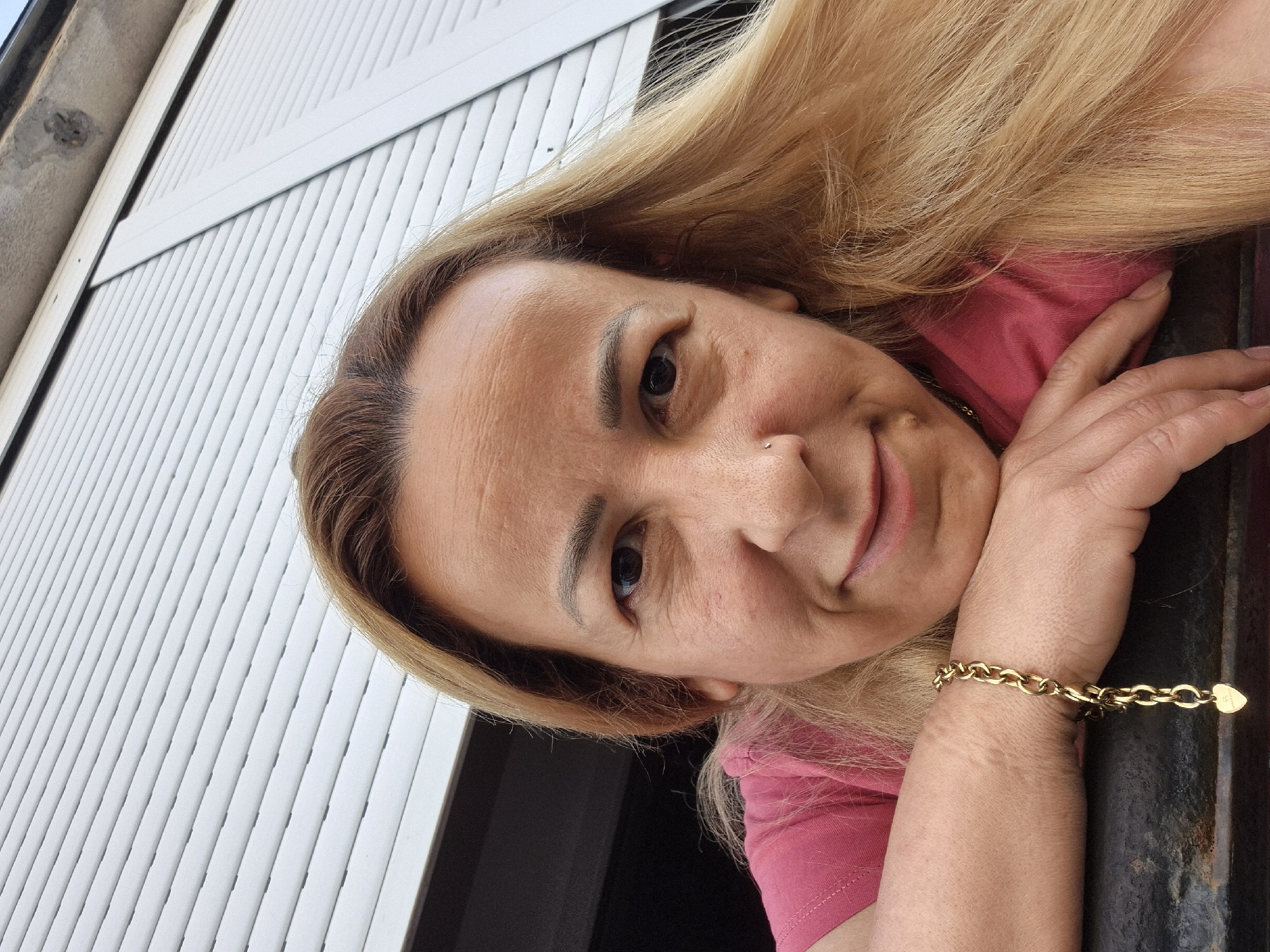 A medium close-up selfie shows a fair-skinned person with blonde hair leaning on a dark surface, their head tilted slightly forward. They are wearing a pink t-shirt. A gold chain bracelet with a heart-shaped charm is visible on their left wrist. The background consists of a white roller shutter and a dark area. The lighting is natural, creating soft shadows on the face. The person appears to be outdoors.