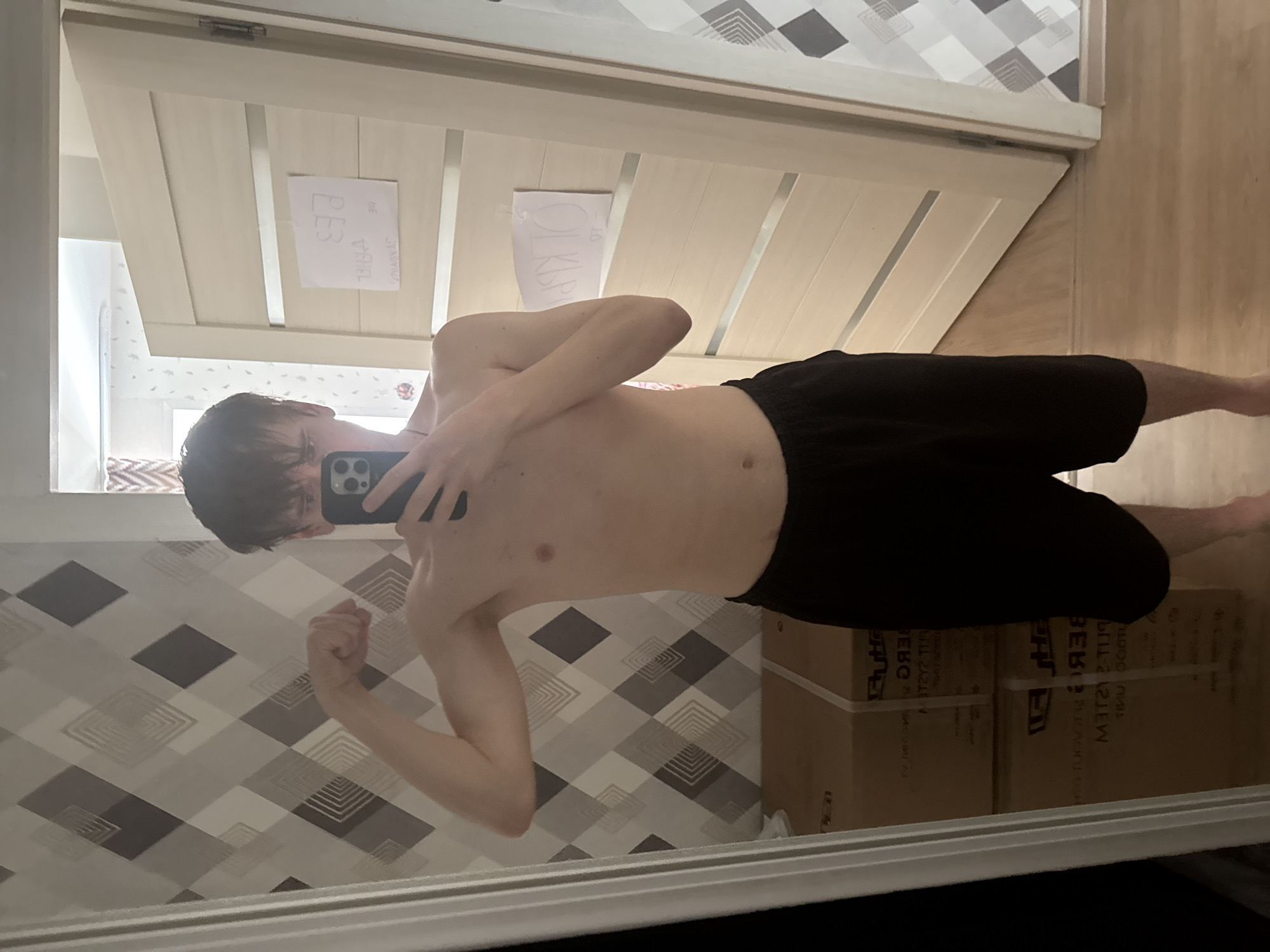 The image shows a young person, appearing to be male, taking a selfie in a mirror. He is shirtless, wearing black shorts, and flexing his bicep. The background suggests an indoor residential setting, possibly a bedroom or hallway. The walls have a geometric diamond pattern wallpaper. There are two doors visible; one is closed and made of light-colored wood with horizontal inlays, and the other is open, revealing a portion of another room. On the closed door, two pieces of paper are taped. One has Cyrillic text written on it, appearing to say "РЕБ" and another word below it, possibly "ТРИГА". The second paper has Cyrillic text that appears to say "ОТКРЫТО" and "97" below it. In the foreground, to the left, are two large cardboard boxes stacked on top of each other. The boxes have text printed on them, also in Cyrillic, which appears to be part of a product name and possibly the brand "RUH / RUZ..." and "SYSTEM / UNIT". The floor is made of light-colored wood planks. The lighting suggests it is daytime. The overall scene is informal and personal, consistent with a home environment. The location of Perm, Russia is inferred from the use of Cyrillic text on the papers and boxes.