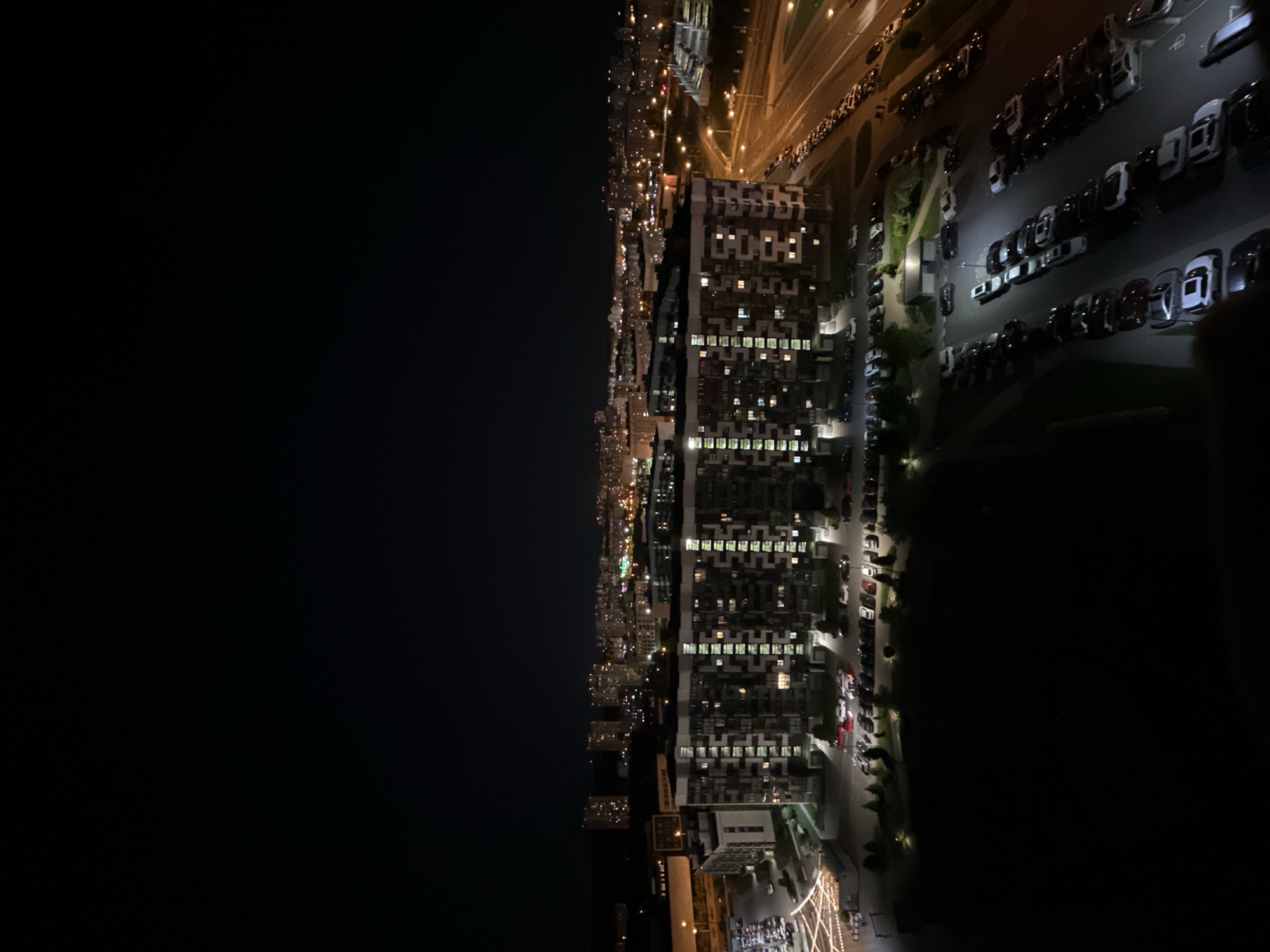 The image depicts an aerial nighttime view of Бараўлянскі сельскі Савет, Belarus. A high-rise residential building with numerous illuminated windows is visible, indicating occupancy. Adjacent to the building is a parking lot filled with parked cars. A city skyline stretches into the distance along one side of the frame. The other side of the frame is mostly dark. Streetlights and car headlights illuminate the streets and parking areas. The environment is urban and illuminated artificially due to the nighttime setting.