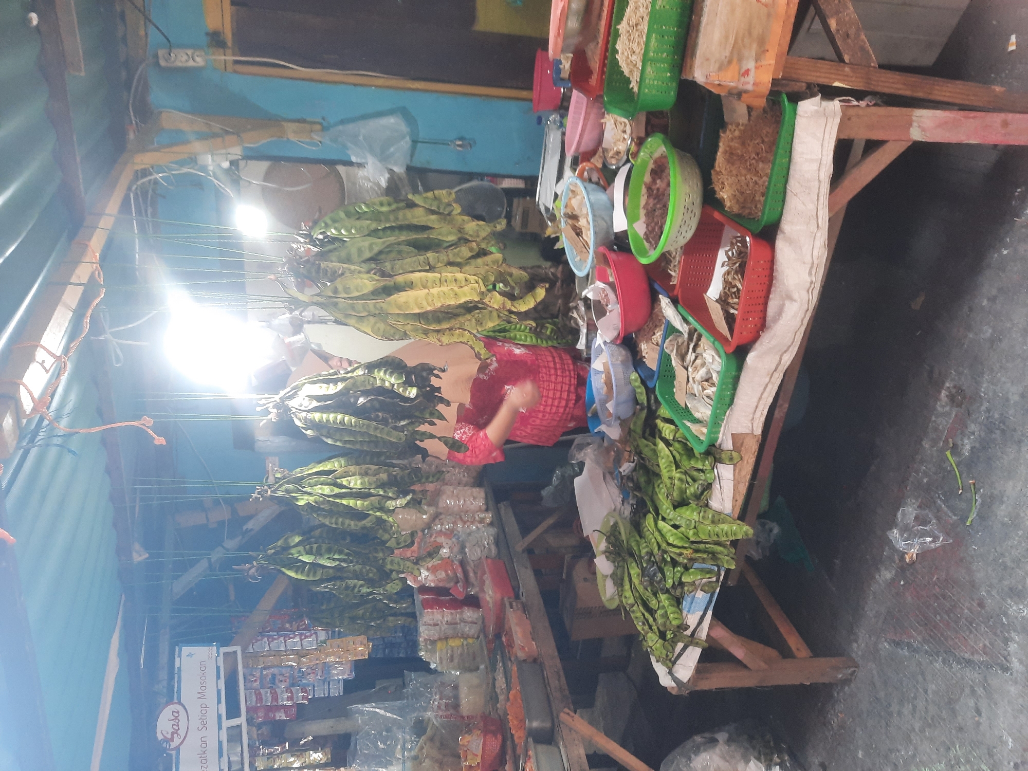 This image depicts a stall in a market, likely located in North Jakarta, Indonesia, with goods displayed on tables and hanging from the ceiling. A woman in a red and white patterned top is visible behind the stall, appearing to be attending to the merchandise. Hanging from the ceiling are several bunches of green legumes, which appear to be petai, a type of stinky bean common in Southeast Asia. In front of the stall, various food items are arranged in containers. These include what seem to be dried fish, anchovies, and fried shallots, presented in colorful plastic baskets. The stall itself is made of wood and is covered by a corrugated metal roof. The environment appears somewhat cluttered, with goods, packaging, and some debris on the floor. The lighting suggests it is either early morning or late evening, or the stall is lit by artificial light sources, including a bright light bulb hanging from the ceiling. The overall impression is that of a functional, local market stall, possibly in a semi-covered outdoor area.
