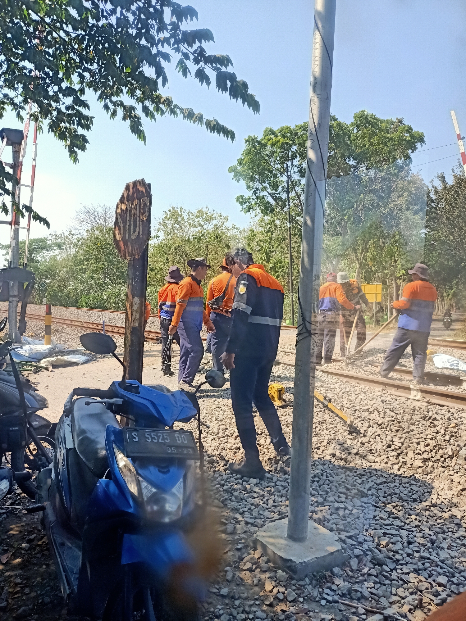 In the city of Banjarjo, Indonesia, a group of workers in orange and blue uniforms are engaged in maintenance work on a railway track. A blue motorcycle is parked in the foreground, its license plate partially visible as "S 5525 DO." In the background, a "STOP" sign is affixed to a post, and trees line the area. The sky is clear and blue, indicating a sunny day. The scene appears to be a railway crossing or a section of track undergoing repair.