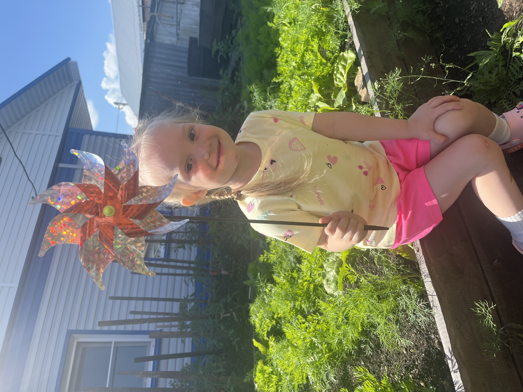 A young girl with blonde pigtails smiles at the camera, holding a colorful pinwheel in her left hand. She is wearing a yellow t-shirt with small fruit and heart graphics, and bright pink shorts. She sits on the edge of a raised garden bed filled with lush green plants, appearing to be in a backyard or garden setting. Her knees are pulled up towards her chest, and her right hand rests on her left knee. The background shows a blue and white building with a ribbed siding, and a glimpse of a clear blue sky with wispy clouds. The sunlight suggests it is daytime, likely a sunny day. The overall scene conveys a sense of joy and outdoor play.