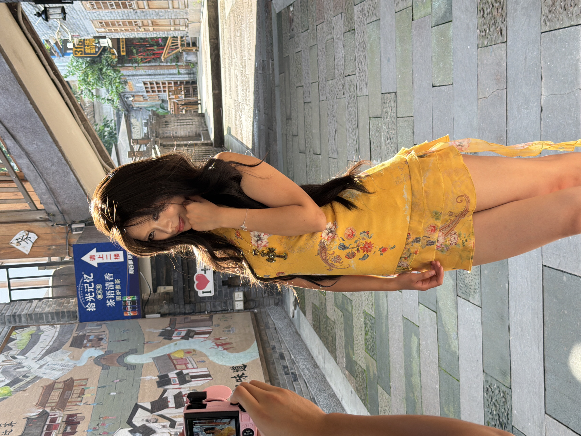 A young woman is posing for a photograph in an alleyway in the Yuzhong District of China. The alley is paved with stone slabs and lined with brick buildings. On the left side of the alley, there is a mural depicting traditional Chinese architecture and people. A pink digital camera is being held up to take the photo. The woman is wearing a yellow floral dress and a gold necklace. She is standing with her hand on her chin, looking off to the side. The sun is shining, casting shadows on the ground.