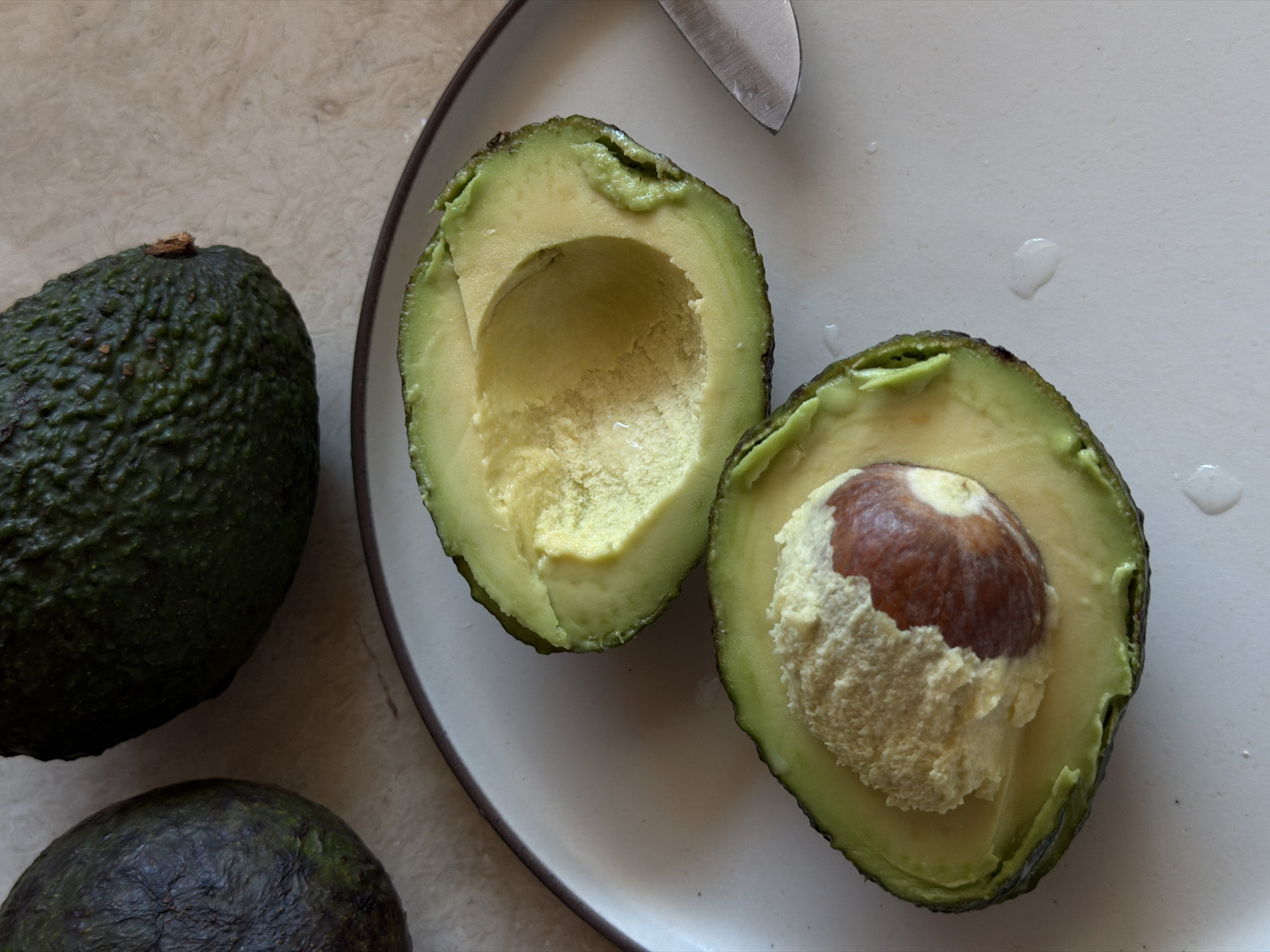 An overhead shot shows two whole avocados and a single avocado halved on a white plate. The halved avocado is in the foreground, with one half showing the pit and the other half showing the empty cavity. The plate is on a textured surface, possibly a countertop, with a knife to the right of the plate. This image does not contain any information about the city of Mengwi, Indonesia.
