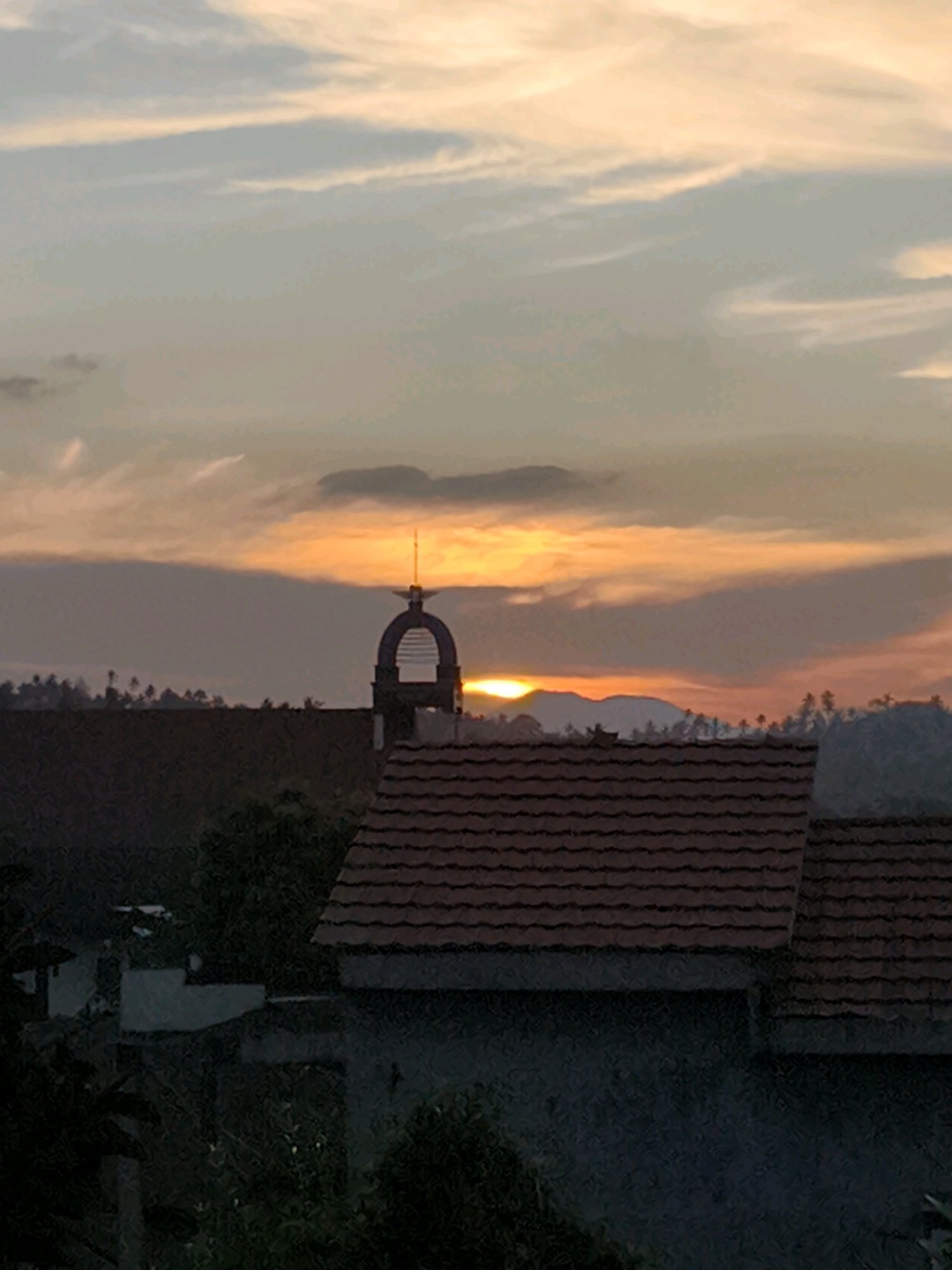 The image depicts a sunset over Tombatu Dua, Indonesia, with the sky painted in hues of orange, yellow, and soft pink, streaked with wispy clouds. In the mid-ground, a distinct bell tower with a pointed top stands out against the horizon. Silhouetted trees line the distant hills. In the foreground, the dark roofs of buildings are visible, one with red tiles and another darker, flatter roof. The overall scene is tranquil, capturing the end of the day.