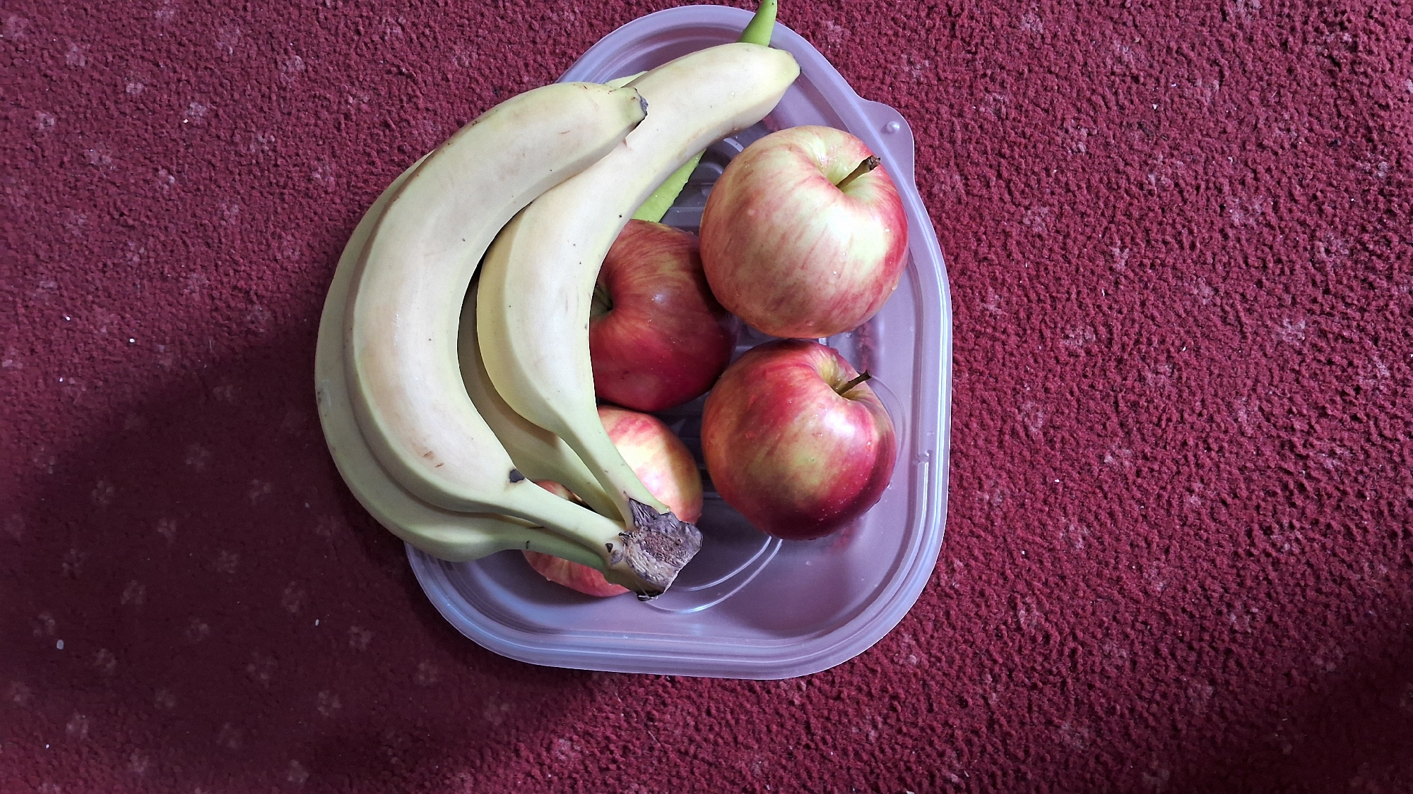 A clear, top-down medium shot shows a clear plastic food container filled with five pieces of fruit: a bunch of three yellow bananas and four red-yellow apples. The container is placed on a maroon carpet. The bananas are positioned over the apples. The apples are arranged in a way that shows their round bodies. Some of the apples have red and yellow stripes. The scene is well-lit, and the colors are vibrant. There are no people visible in the image. The context of Varna, Bulgaria is not depicted in this image.