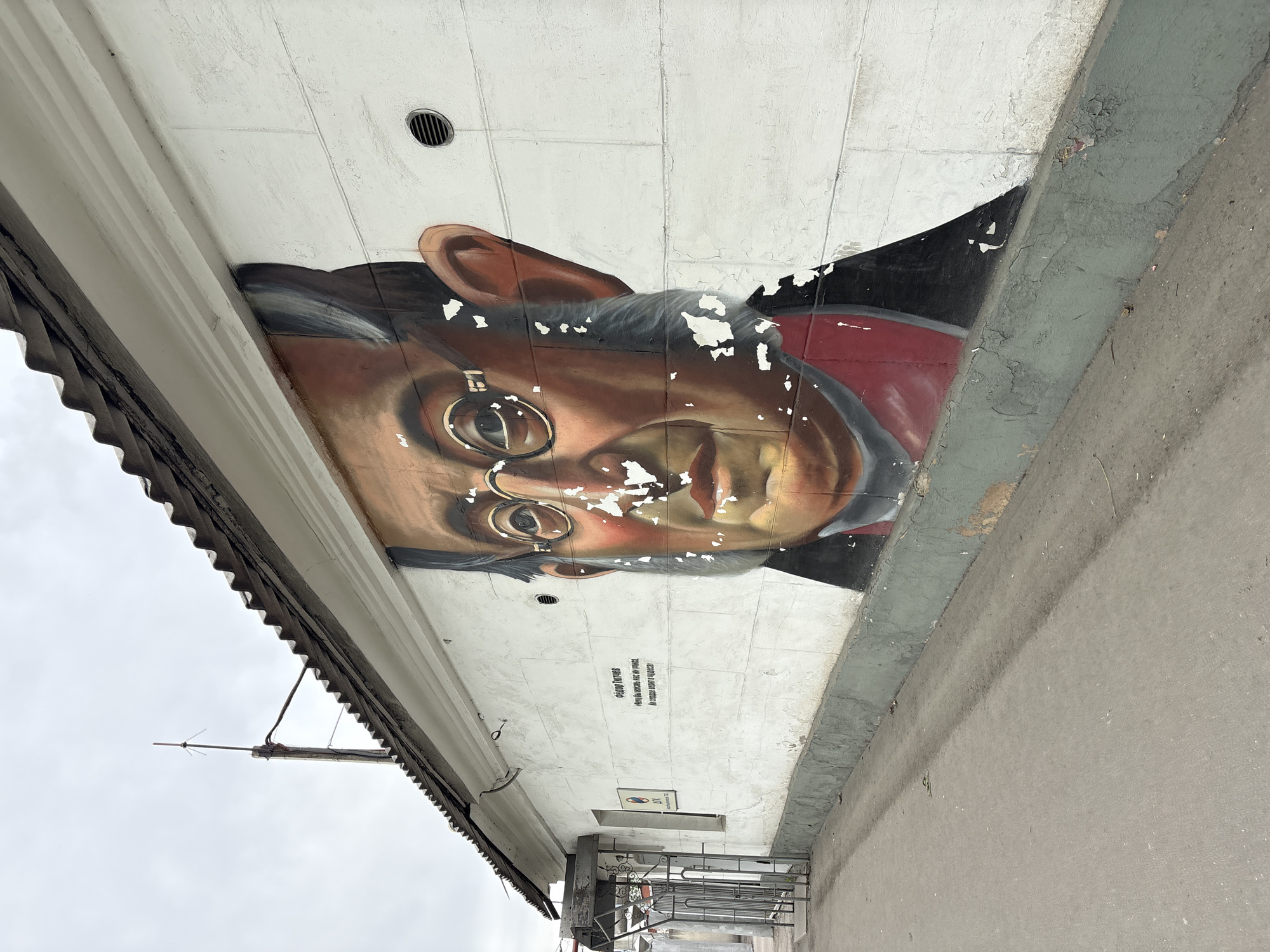 The image shows a mural of Sholom Aleichem, a Yiddish author and playwright, painted on the underside of a building overhang. The mural is located in Birobidzhan, Russia. The building's overhang has a corrugated metal roof on the left side and a grey concrete ledge on the right. The mural depicts Aleichem's face, wearing round spectacles. Above and below the mural, the painted wall is peeling. Below the mural, there is writing in a Cyrillic script and a small sign. Below the overhang, there is metal scaffolding. The sky above appears overcast.