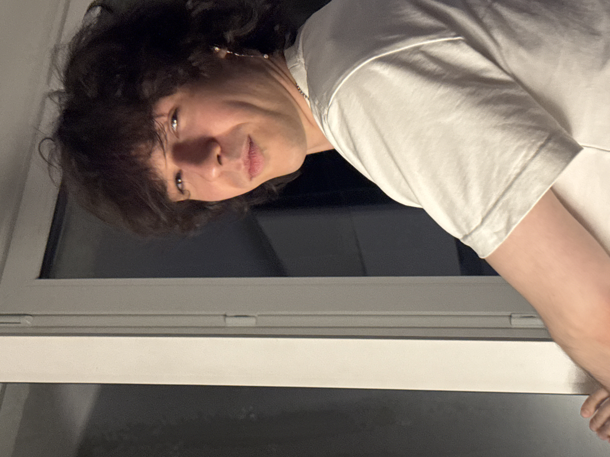 The image shows a young man with dark, curly hair, looking over his shoulder towards the camera. He is wearing a white t-shirt. The man appears to be indoors, standing next to a window frame. The lighting suggests it is nighttime or an indoor setting with artificial light. There is a dark background behind him, and a white vertical element, likely a window frame or door frame, to his left. The man's right ear is adorned with a long, dangling earring. The image is a medium shot, focusing on the upper body and face of the individual.