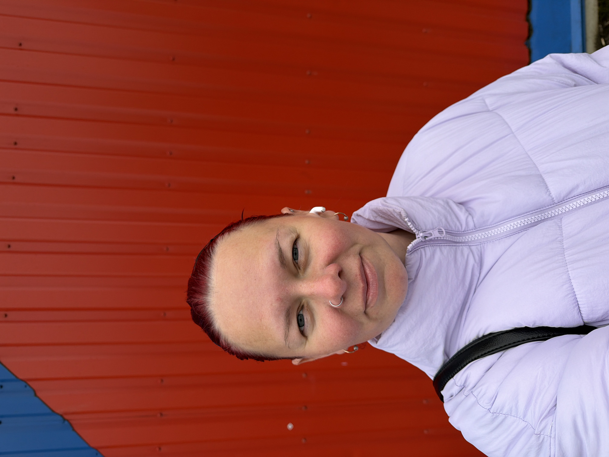A medium, close-up shot shows a light-skinned woman with short, red hair, wearing a light purple puffer jacket. She has blue eyes and a silver nose ring. Behind her is a wall composed of red and blue corrugated metal panels. Based on the location, the image was taken in Chaikovsky, Russia.