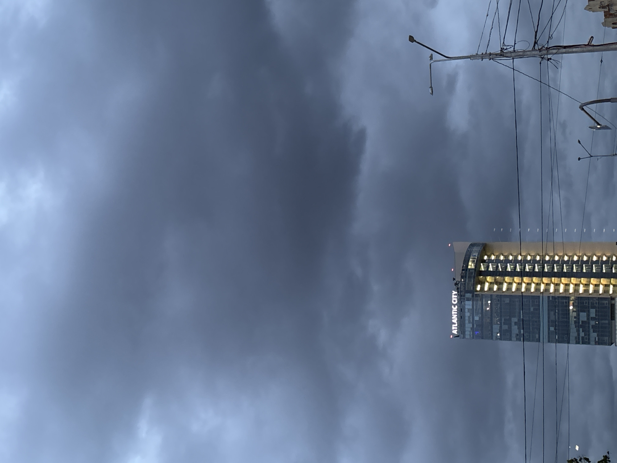 The image shows a tall building with the words "ATLANTIC CITY" vertically displayed on its side. The building has many illuminated windows. Above the building, there are numerous power lines and a streetlight. The sky is overcast with dark, grey clouds. The location is likely in the city of Saint Petersburg, Russia.