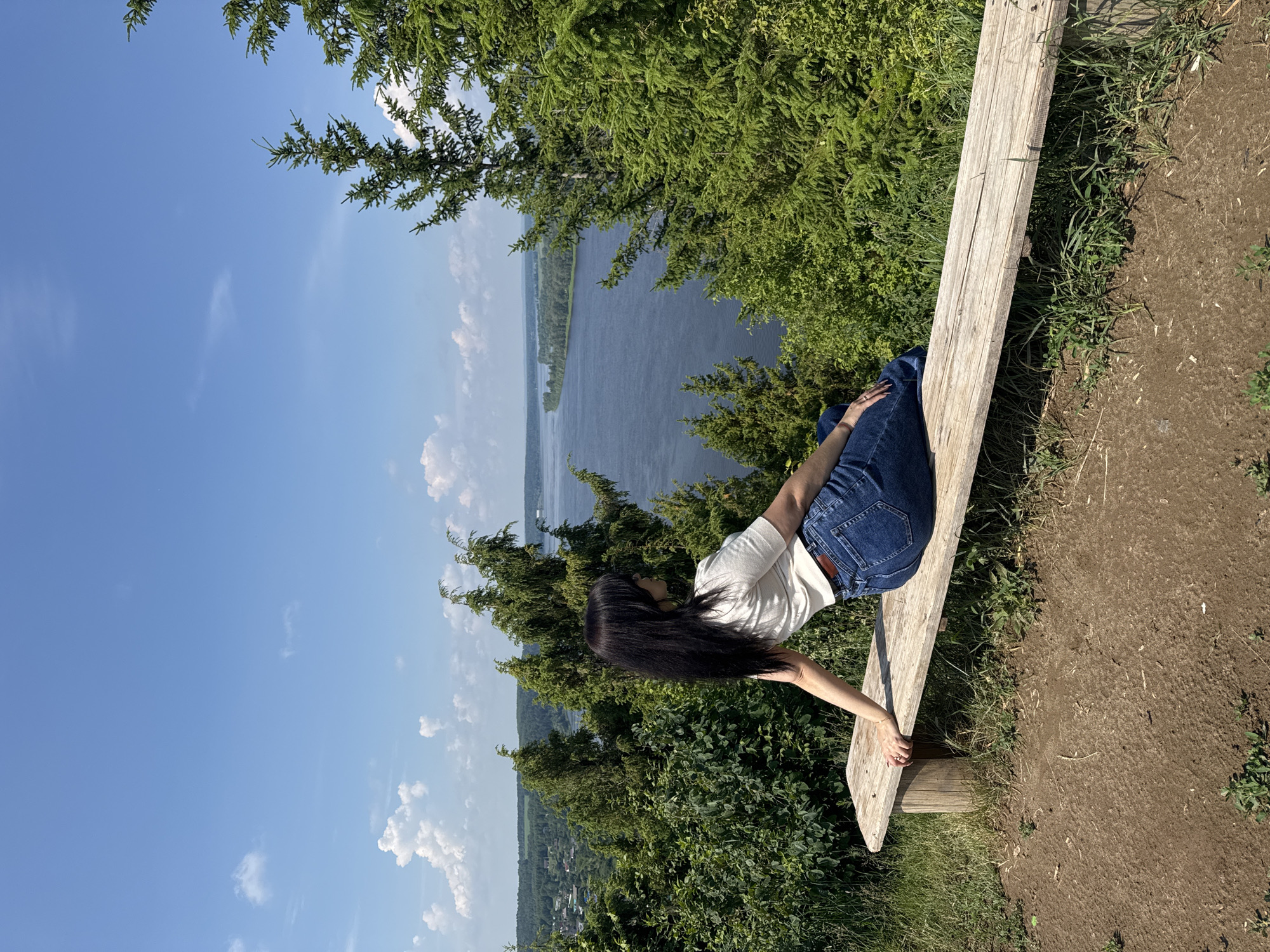 A young woman with long black hair is sitting on a wooden bench, looking out at a wide river. She is wearing a white t-shirt and blue jeans. The bench is positioned on a grassy embankment overlooking the river, with trees and vegetation surrounding the area. In the distance, a town can be seen on the opposite bank of the river. The sky is clear and blue with scattered white clouds. The location is identified as Olkhovka, Russia.