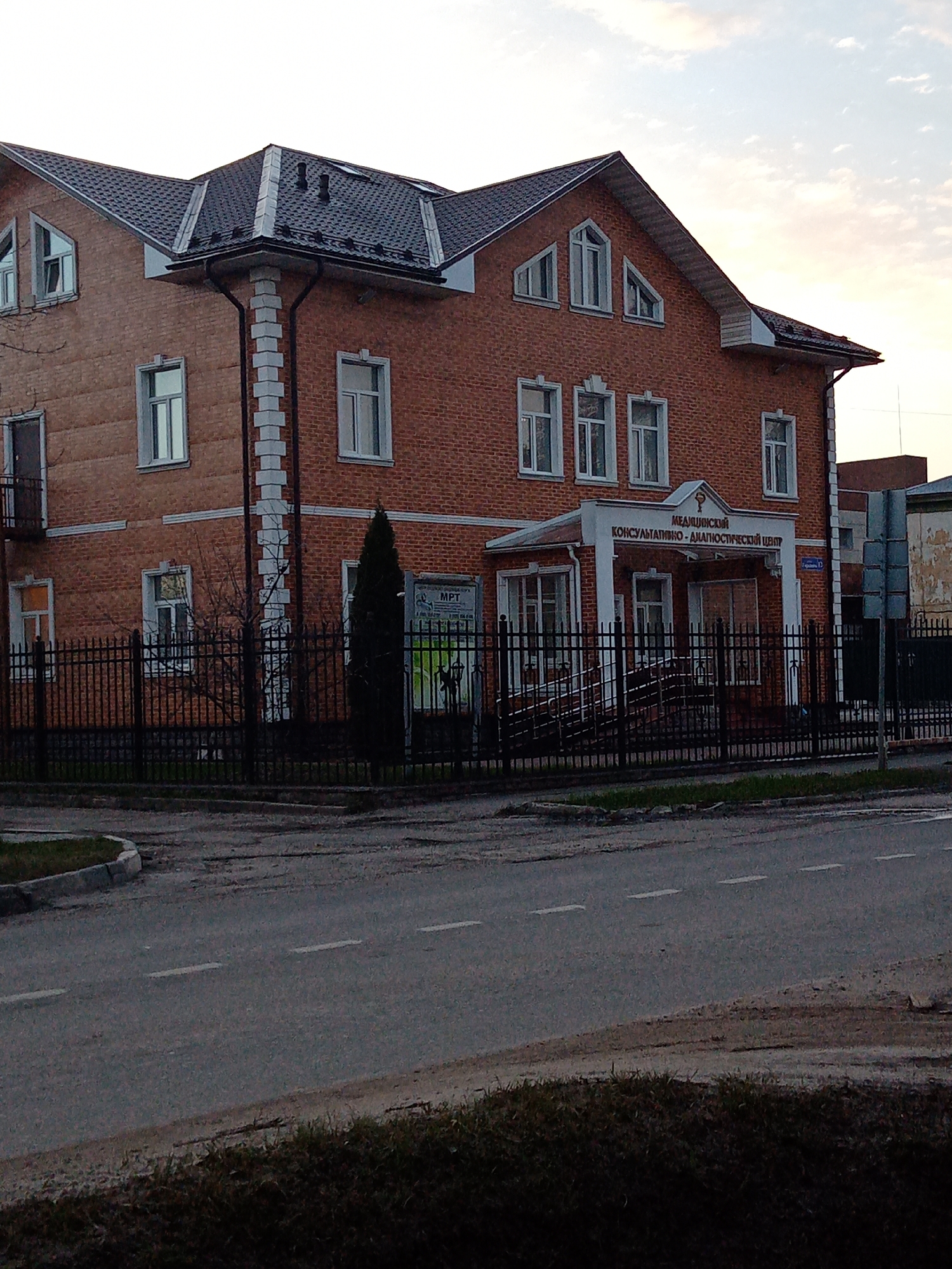 The image depicts a two-story brick building with a dark gray roof, identified by a sign in Russian as a medical consultative-diagnostic center. The building has white-framed windows and a white portico over the entrance. A black wrought-iron fence surrounds the property. A narrow tree stands near a sign that reads "MPT." The building is located at Pionerskaya Street 19A in Elektrostal, Russia. The scene is viewed from across the street, with a portion of the road visible in the foreground. The sky is overcast, suggesting early morning or late afternoon lighting. No people are visible in the image.