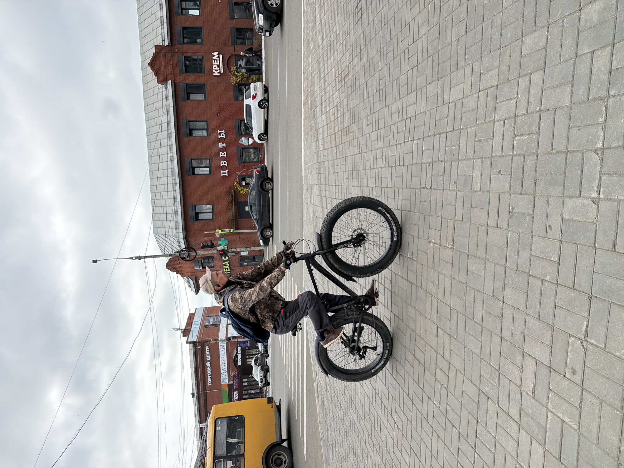 A man rides a fat-tire bicycle on a paved sidewalk in front of brick buildings in Votkinsk, Russia. The man is wearing a camouflage jacket, dark pants, and a light-colored baseball cap. He has a blue backpack on. The buildings have signs in Russian, including "ТОРГОВЫЙ ЦЕНТР" (Shopping Center), "ЦВЕТЫ" (Flowers), and "КРЕМ" (Cream). A yellow bus is visible on the left side of the image, and several cars are parked or driving on the street in the background. The sky is overcast. The sidewalk is made of gray interlocking pavers.