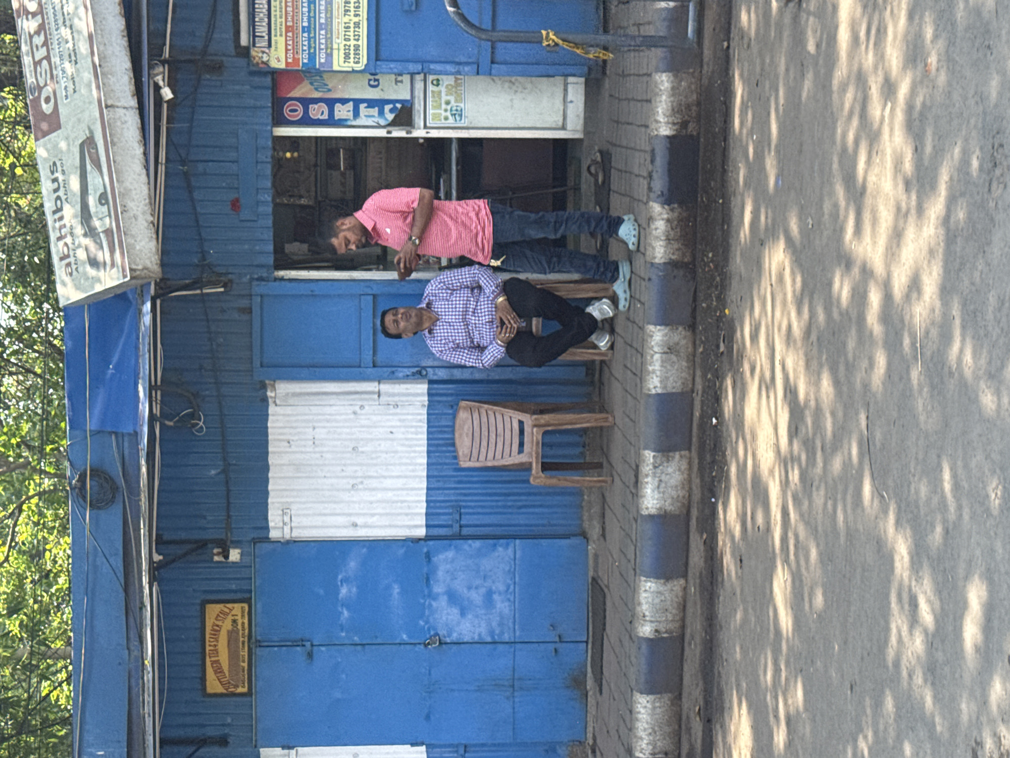 A street scene in Kolkata, India. Two men are present outside a blue-painted tea and snack stall. One man is wearing a pink shirt and dark jeans, the other a light purple checked shirt and black pants, sitting on a plastic chair. The stall’s signage includes the names "abhibus," "OSRTC," and "NILAMADHABA," indicating bus travel services. The stall also provides contact numbers and mentions cities such as Kolkata and Bhubaneswar, potentially indicating a bus route. The street is paved and shows shadows of foliage. A sign reading "TEA & SNACK STALL" is visible.