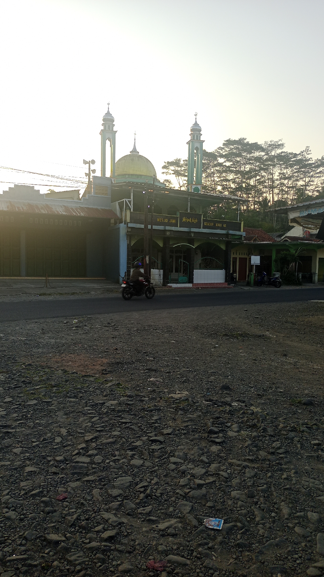 The image captures a daytime scene in the city of Karyamandala, Indonesia, with a mosque as the central focus. The mosque, identifiable by its green minarets and golden dome, is situated along a paved road. In the foreground, the road is flanked by a gravelly area. A person is seen riding a motorcycle on the road in front of the mosque. The background features a line of trees silhouetted against a bright sky, suggesting either early morning or late afternoon light. The environment appears to be a mix of urban and natural elements, with buildings and a mosque alongside a natural tree line.