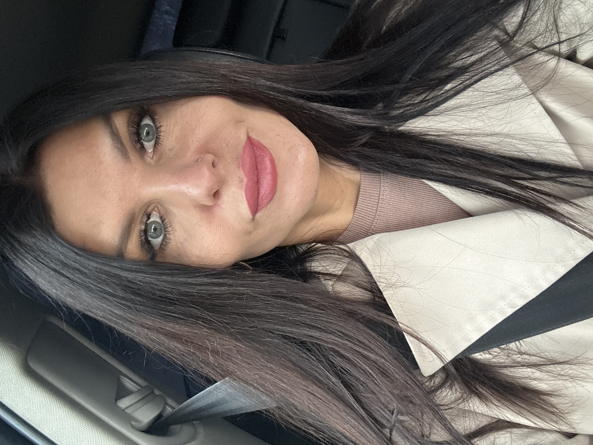 A woman with long, dark hair and light eyes is in a car, presumably on the passenger side as indicated by the seatbelt. She is wearing a beige coat over a pink sweater. The interior of the car is visible, including a dark headrest. The photo shows a close-up of the woman's face, angled slightly. The location is not discernible from the image and there is no information available to suggest it is located in Chaikovsky, Russia.