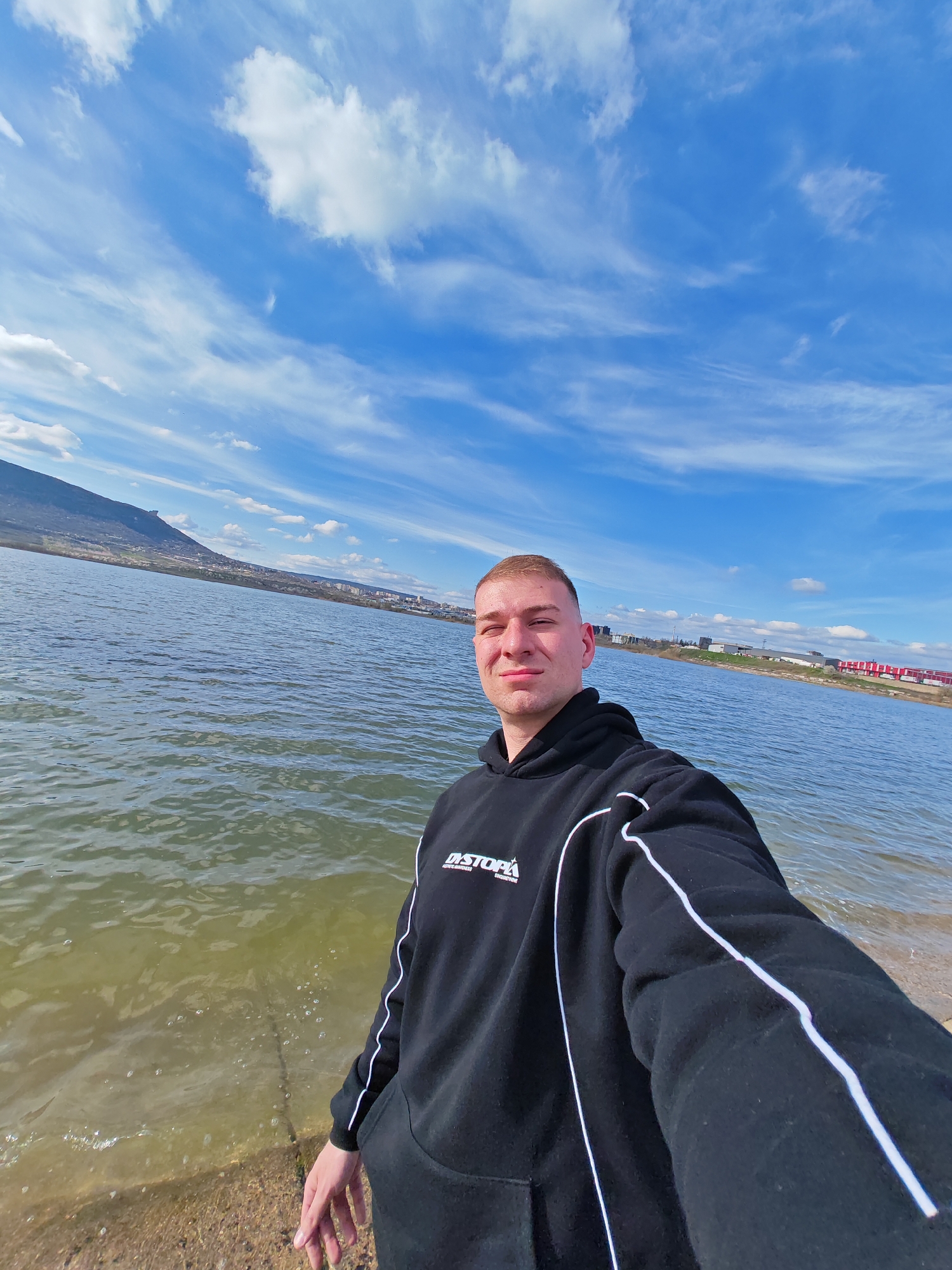 The image shows a man taking a selfie at the shore of a body of water, likely a lake or reservoir, on a sunny day with a blue sky and scattered clouds. The man is wearing a black hooded sweatshirt with white stripes along the sleeves. In the background, there's a mountain range with a settlement on the left side, and on the right are some buildings. Based on the background, it appears to be the Shumen Lake in Shumen, Bulgaria.