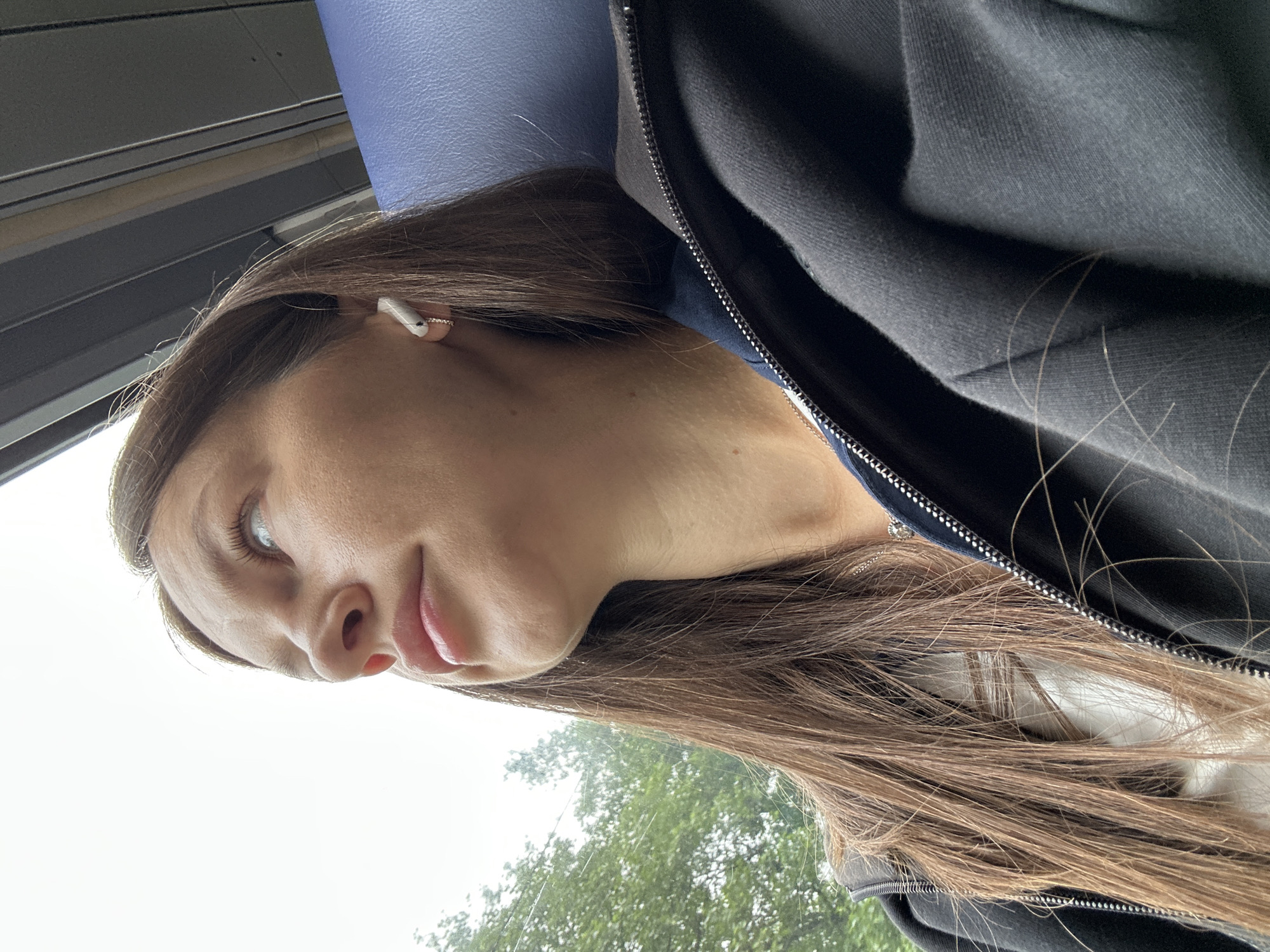 This tilted, close-up portrait features a young woman looking upwards and to her right, conveying a thoughtful or observant mood. She has long, straight brown hair that cascades over her shoulders and a dark, zippered jacket, possibly made of fleece or cotton. A white earbud is visible in her right ear, along with a small, delicate golden hoop earring. A silver necklace chain can be seen peeking out from beneath her jacket near her neck. Her light-colored eyes are directed away from the viewer, and her lips are softly parted in a subtle smile or pensive expression.
The setting appears to be inside a vehicle, possibly a bus or train, given the light blue, textured surface behind her head, which resembles a seat's headrest. Through a window in the background, a bright, overcast sky and the tops of green trees are visible, suggesting it is daytime in Stade, Germany. The natural light illuminates her face softly. No text is visible in the image.