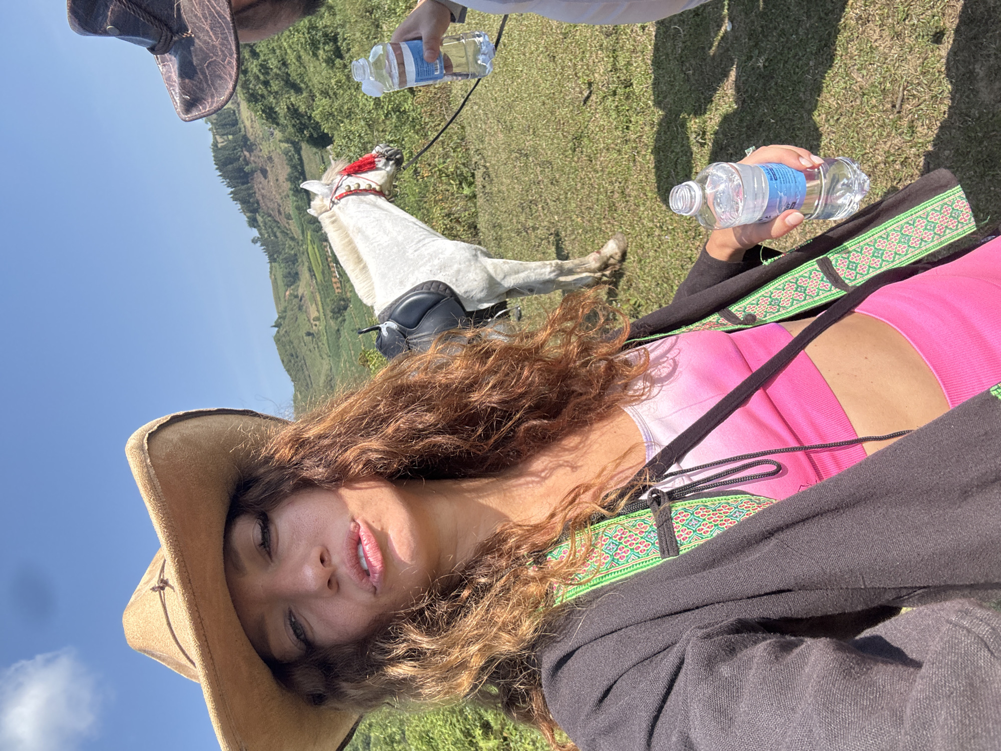 A daytime, tilted, selfie-style shot outdoors features a person wearing a brown cowboy hat and a pink top, with a dark jacket partially visible. The subject has long, curly brown hair and is holding a plastic water bottle in their right hand. To the left, a white horse with a black saddle and a red-trimmed headstall stands on a grassy hill. Another hand holding a water bottle is visible near the horse's head. The background shows a hilly landscape under a clear blue sky with a few clouds. The location is Bản Phố, Vietnam.