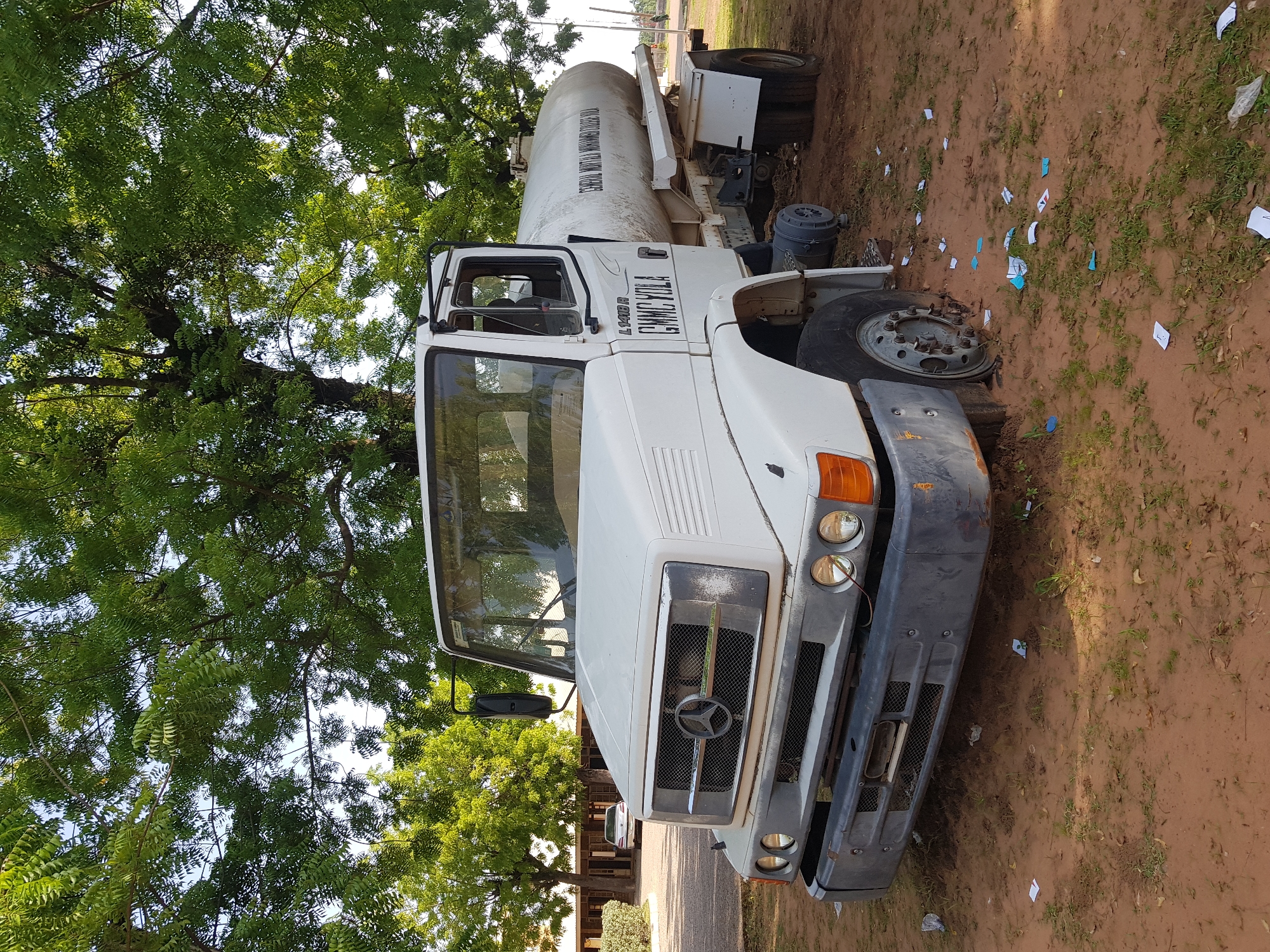 A white Mercedes-Benz tanker truck is parked on a dirt road under a large tree with dense green foliage. The truck has a white cab and a large white cylindrical tank mounted on its chassis. The truck's grille features the Mercedes-Benz logo. The bumper is a dark grey. The truck appears to be stationary. Scattered on the ground around the truck are pieces of paper and some vegetation. The image is taken during daylight hours. The overall scene suggests an outdoor setting in a rural or semi-rural area, possibly in Jimeta, Nigeria, given the location context. There are no people visible in the image. The truck has text on its side that reads "GMAC YOLA" and on the tank the text "CHEMICAL WORKS YOLA".