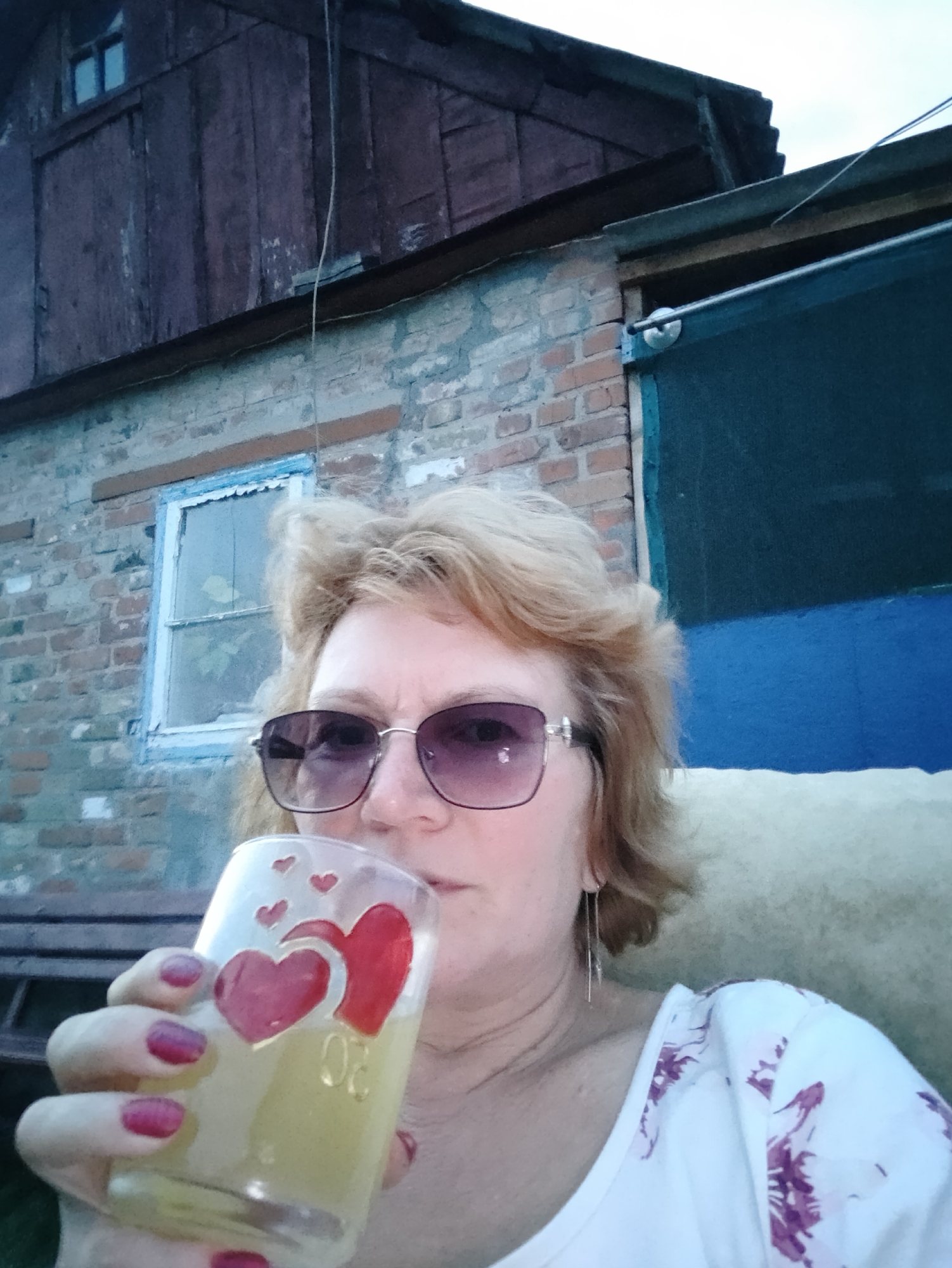A medium shot captures a woman with blonde hair and sunglasses holding a glass with red hearts, as she drinks from it. She is seated in front of a weathered brick building. The building appears to be an outbuilding or shed with a wooden upper section and a blue tarp or door on the right. The overall scene suggests an outdoor setting, possibly in the backyard of a residential property in Kremenchuk, Ukraine, given the context.