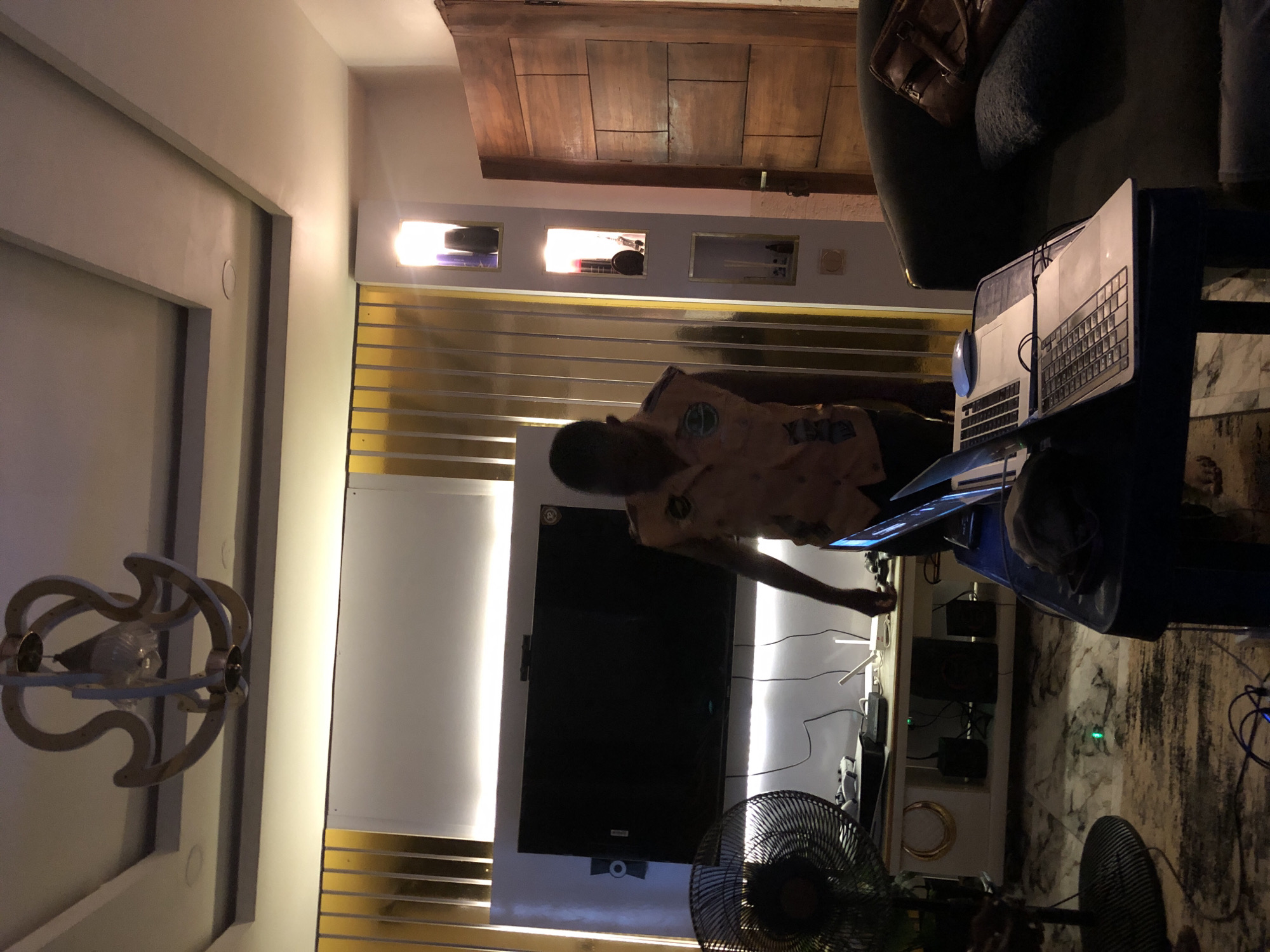 This media file captures an indoor room, likely a living room or entertainment area, rotated 90 degrees counter-clockwise.
A dark-skinned individual, possibly male, wearing a light-colored, patterned shirt, is positioned in the center, reaching towards a large black flat-screen television. Their face is obscured by shadow.
The room features a modern aesthetic with light-colored walls accented by horizontal gold strips and recessed lighting. The television is mounted on a white panel, and below it, on a white console, are several electronic devices and speakers with visible cabling. To the right of the person, on a blue stand, sits a laptop and a desktop computer with a separate keyboard and mouse. Two black electric fans are visible: one partially in the lower left, and another with a circular base in the lower right foreground.
The background includes a heavy, paneled wooden door. Above the TV area, several small, lighted display niches hold various items. A dark sofa or armchair, topped with a brown leather bag, is partially visible near the wooden door. An ornate ceiling light fixture with swirling metallic elements hangs in the upper left. The floor appears to be covered with a patterned rug or large tiles.
The lighting, primarily artificial from various fixtures, suggests an evening or nighttime setting. No discernible text is visible on any objects or surfaces. The scene depicts a functional space, possibly set up for gaming, work, or media consumption.