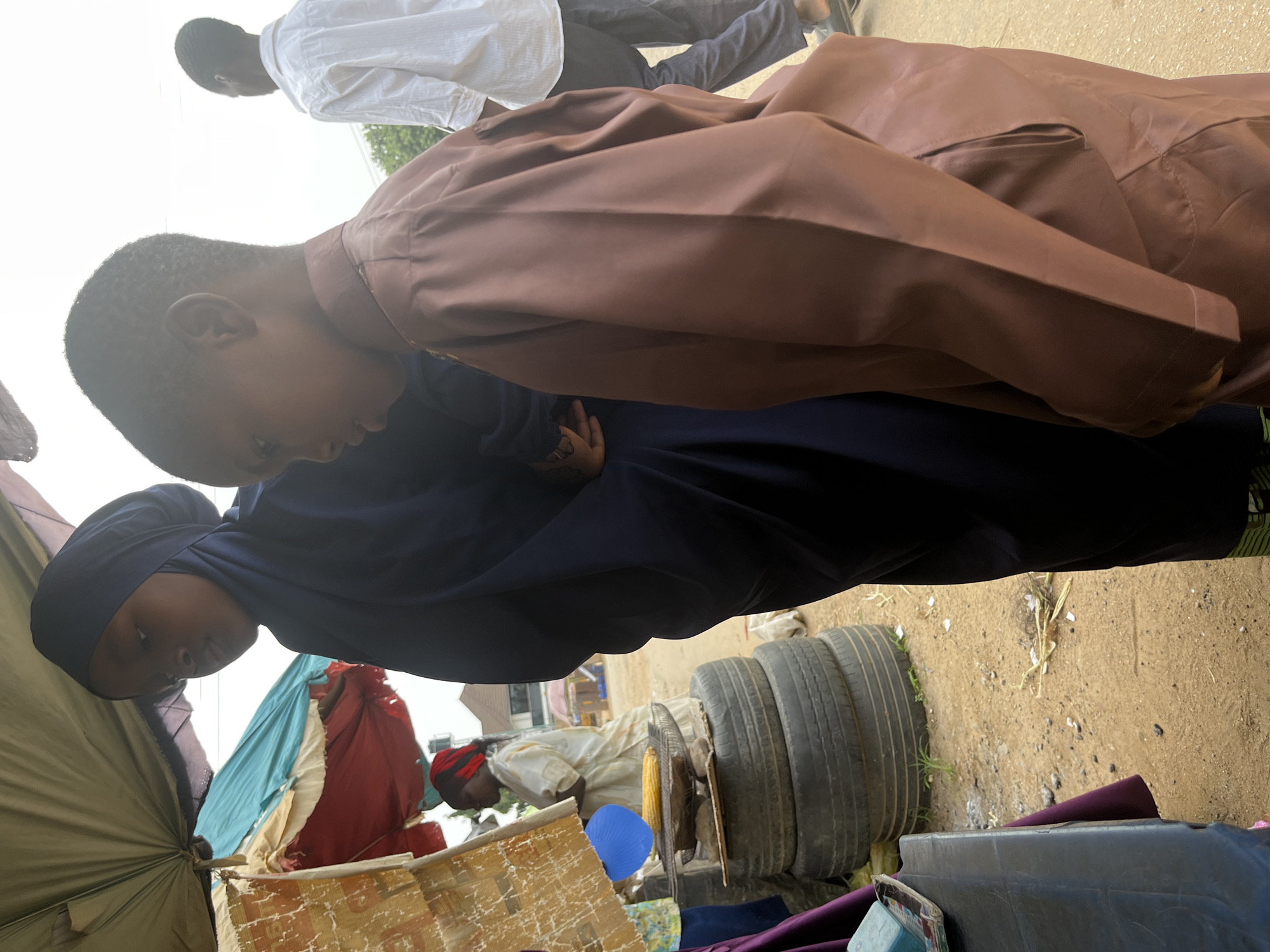 The image depicts an outdoor scene in what appears to be a market in Abuja, Nigeria. In the foreground, a young boy wearing a brown traditional outfit, possibly a kaftan, is captured in profile. To his left, a young girl in a dark blue hijab is also partially visible, facing away from the camera. Behind them, a woman wearing a light-colored garment and a red headscarf is seen tending to a corn cob roasting on a grill, which is supported by a stack of tires. In the background, a man in a white shirt and dark pants walks away from the camera, and other market stalls and structures are faintly visible, suggesting a bustling environment. The ground is sandy and dusty, typical of outdoor markets in the region.