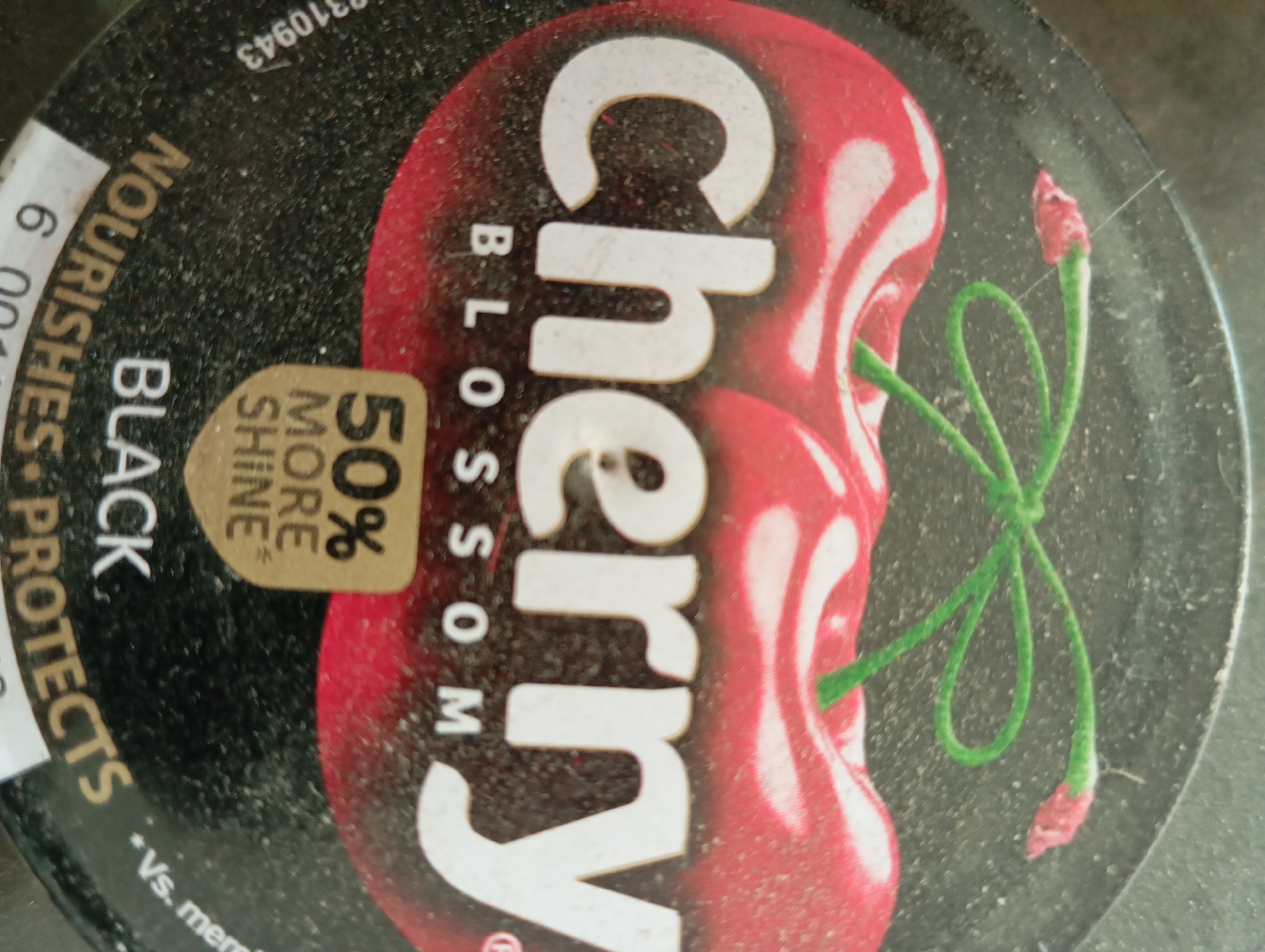 The image is a close-up, slightly angled, top-down view of the lid of a product. The lid is dark and covered in dust. A large graphic of two cherries, one whole and one halved, with green stems and a small blossom is the prominent feature. The words "Cherry Blossom" are printed vertically on the lid in large, white, stylized font, with "Cherry" larger than "Blossom." To the left of the cherries, a white label is partially visible, with the number "6" and text "NOURISHES PROTECTS" printed on it. Below this, the word "BLACK" is visible. A small, tan-colored oval sticker is also on the lid, with the text "50% MORE SHINE" printed on it in black. The bottom-left corner of the lid has a small amount of white text that appears to be an asterisk and "Vs. mer". There are no people or any discernible city elements present in the image.