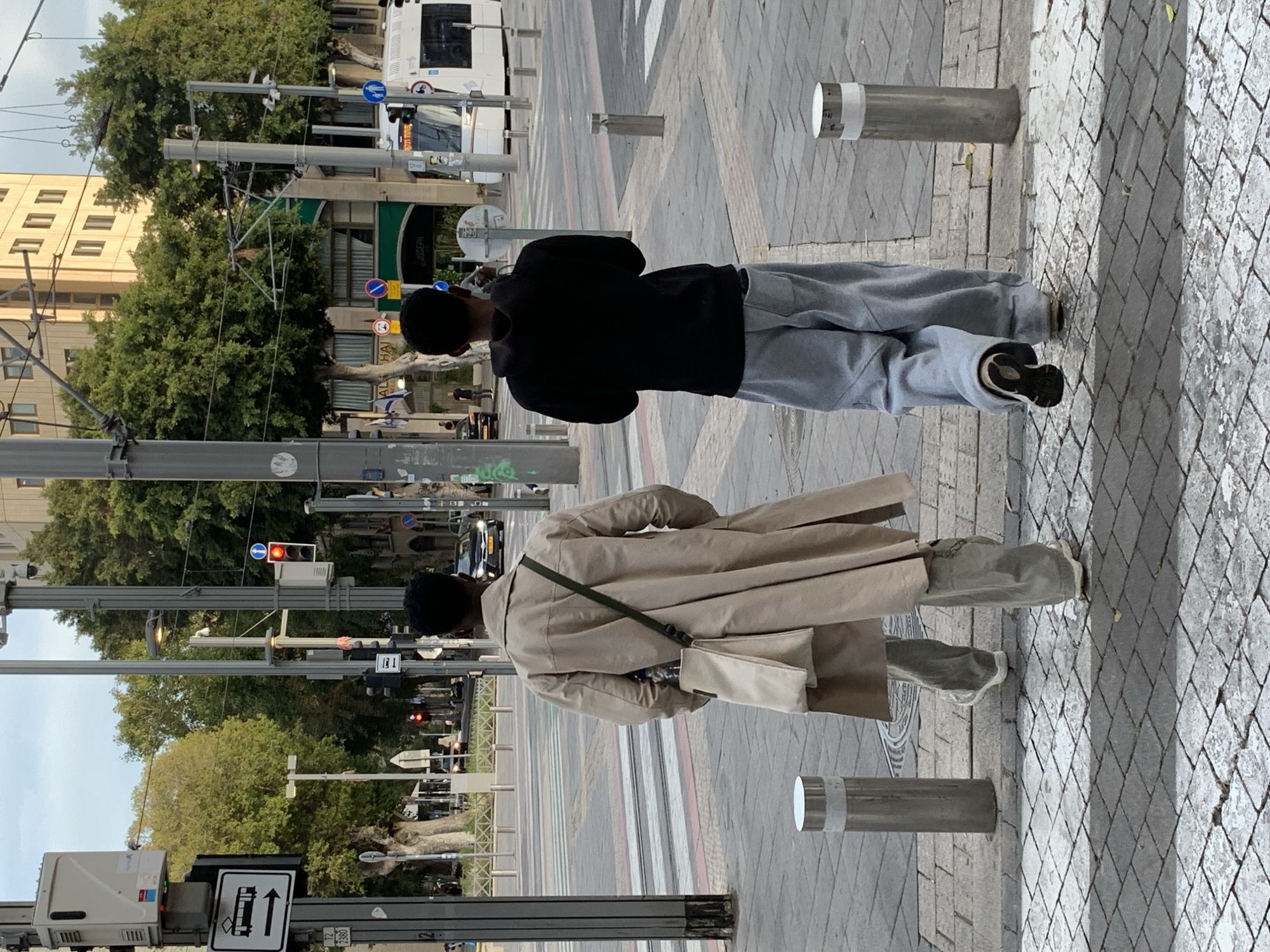 Two individuals are walking on a sidewalk in Tel-Aviv, Israel, as seen from behind. The first individual wears a black hoodie and grey sweatpants. The second individual wears a beige trench coat and carries a beige bag with a green strap. The sidewalk is paved with light grey bricks and bordered by metal bollards. Street lights and overhead tram wires are visible along the street. Trees line the road, and buildings are present in the background. A tram is also visible in the background.