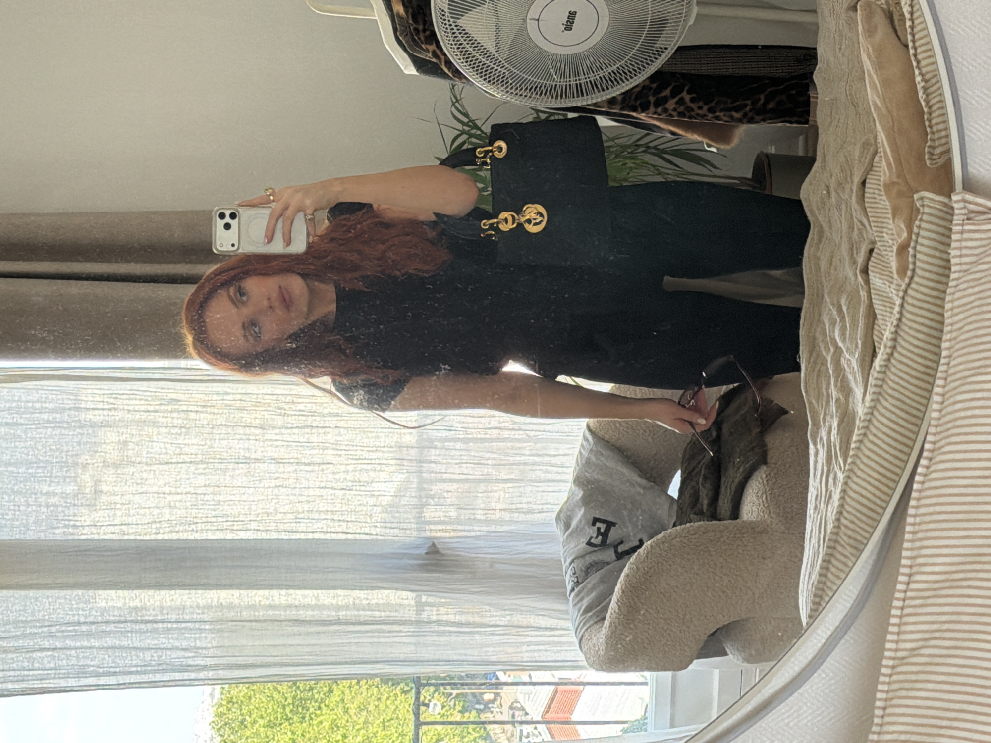 A woman with red hair is taking a selfie in a mirror. She is wearing a black outfit and carrying a black handbag with gold hardware. The mirror reflects a room with curtains, a view of buildings and trees outside the window, a fan, and various items of clothing. Based on the architecture and the view from the window, the location is likely in Paris, France.