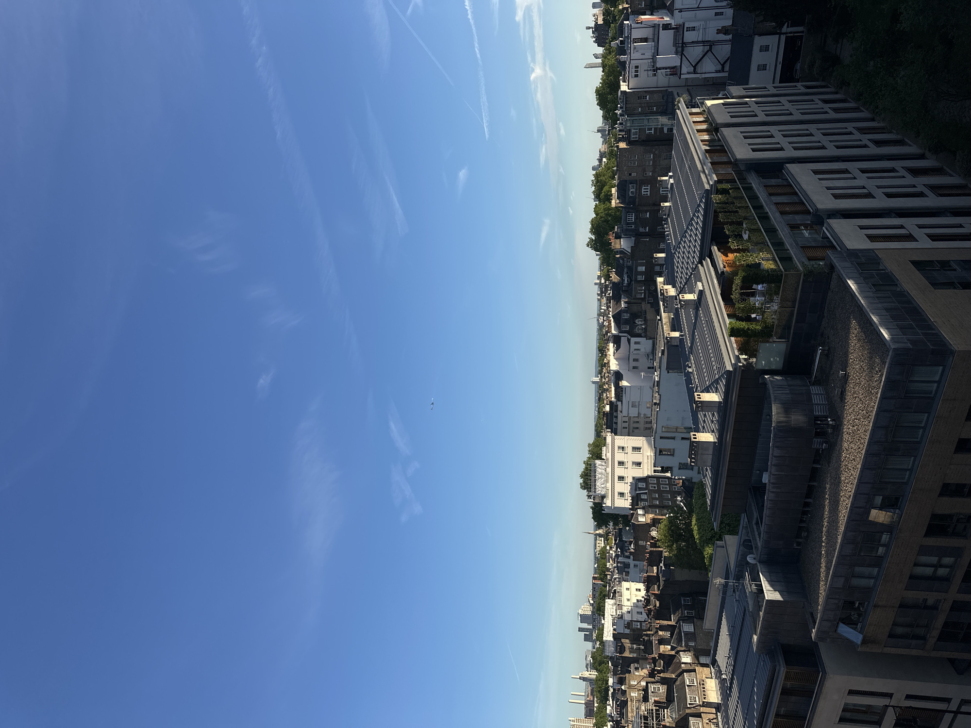 The image shows a cityscape under a bright blue sky with faint contrails. The buildings are a mix of old and modern architecture, with some greenery visible. The location is likely in London, United Kingdom, based on the architectural style and overall urban landscape. There are no people visible in the scene.