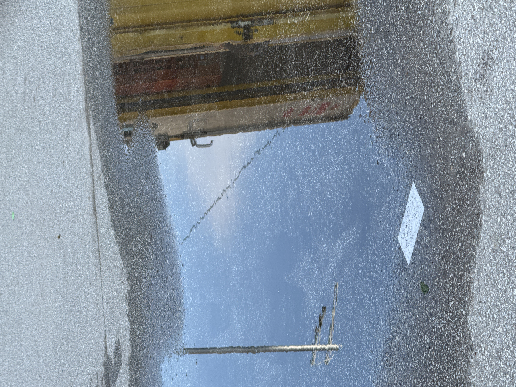 A vertical, medium-angle shot captures a puddle on a grey asphalt surface. The puddle reflects the sky, a pole with horizontal attachments, and a building with vertical yellow and red panels. A white rectangular marking is visible on the asphalt to the left of the building's reflection. A small green leaf rests on the asphalt near the white marking. The scene appears to be an outdoor urban environment.