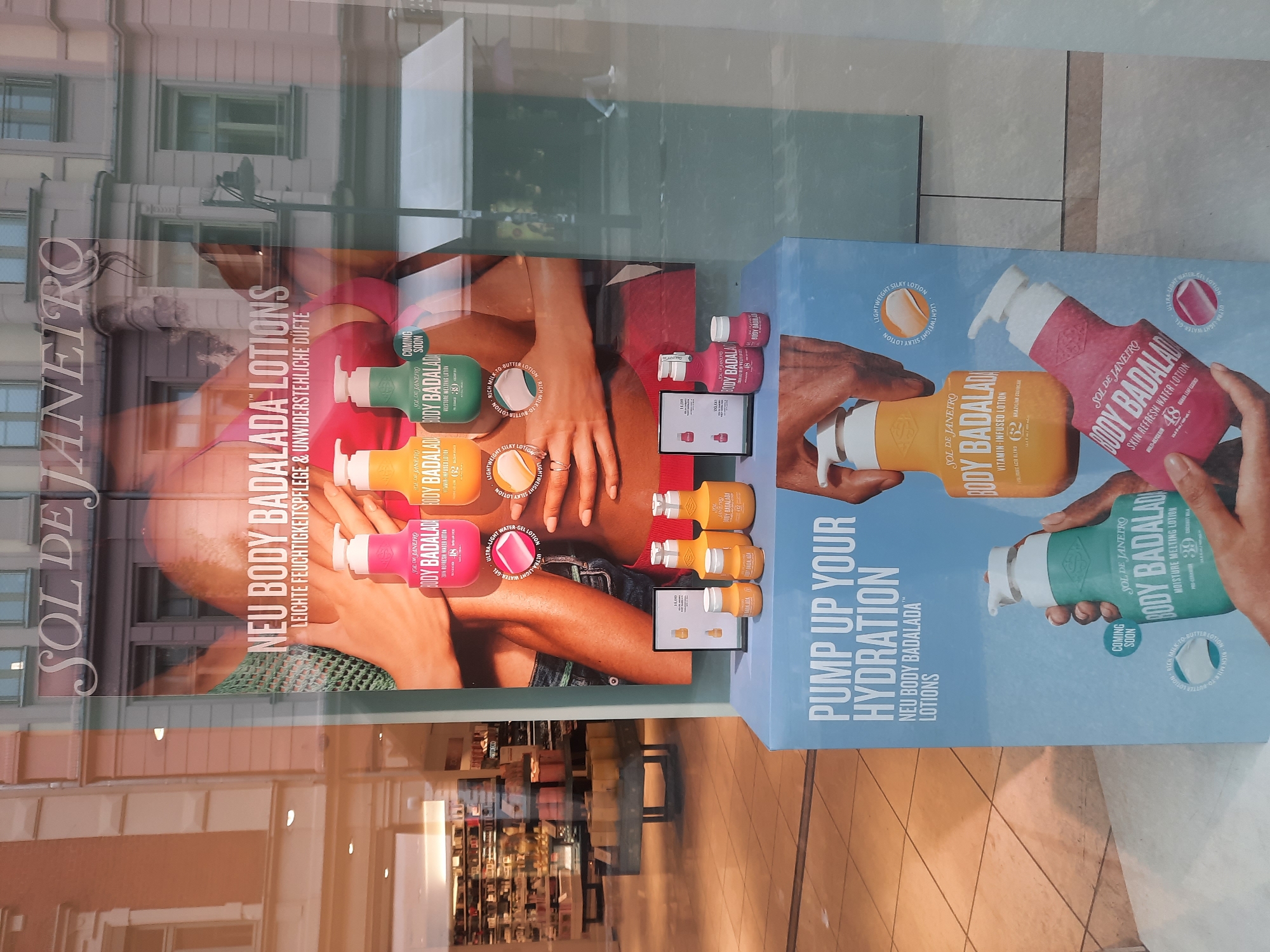 This is a full-shot, eye-level, interior photograph of a storefront window display, likely taken during the daytime. The display showcases the brand "SOL DE JANEIRO" and its "BODY BADALADA" lotions. The main advertising poster features a close-up of a woman's torso, with a focus on her hands holding various colored bottles of the lotion. Text on the poster reads "NEU BODY BADALADA LOTIONS LEICHTE FEUCHTIGKEITSPFLEGE & UNWIDERSTEHLICHE DUFTE". Below this, a pedestal display features several bottles of the lotions in different sizes, including a digital display showing one of the products. Further down, another large poster depicts hands holding bottles of the lotion against a light blue background with the text "PUMP UP YOUR HYDRATION NEU BODY BADALADA LOTIONS". The storefront glass reflects the exterior, including architectural details of buildings across the street, suggesting an urban setting. The reflection shows windows and facades of buildings, consistent with a city like Rosenheim, Germany. The floor inside the store, visible through the window, is tiled. The overall impression is a promotional display for beauty products in a retail environment.
