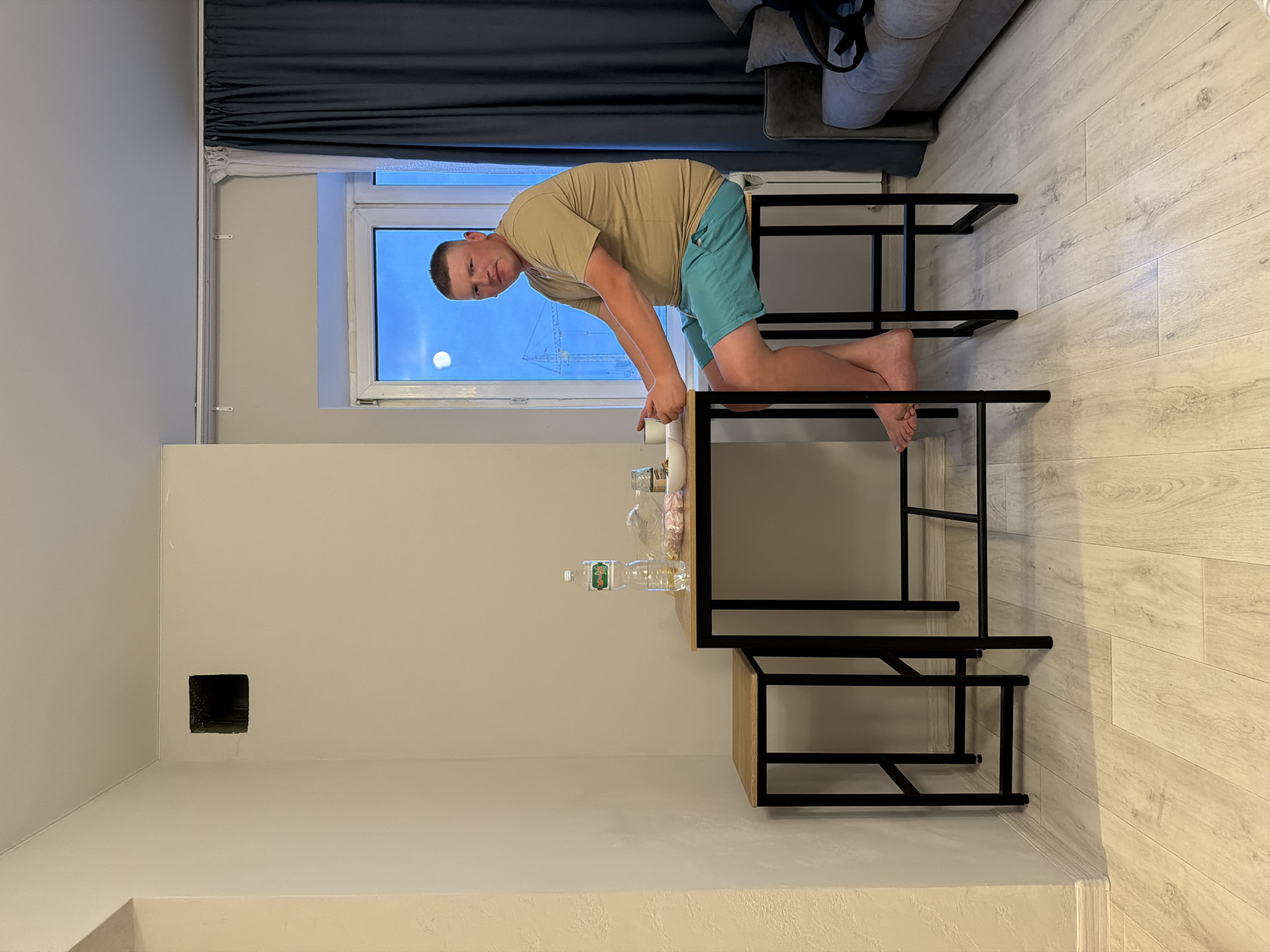 A person is kneeling on a multi-tiered metal and wood table in an indoor setting. The person is wearing a tan t-shirt and turquoise shorts. They are leaning forward with their hands near the top tier of the table. The table has various items on it, including a clear bottle of water and a white container. A window is in the background, showing a view of Naberezhnye Chelny, Russia, with construction cranes visible in the distance. A dark curtain hangs to the left of the window. The room has light-colored walls and wood-look flooring. A dark, square opening is in the wall to the left. A gray couch is in the upper-right corner of the image.
