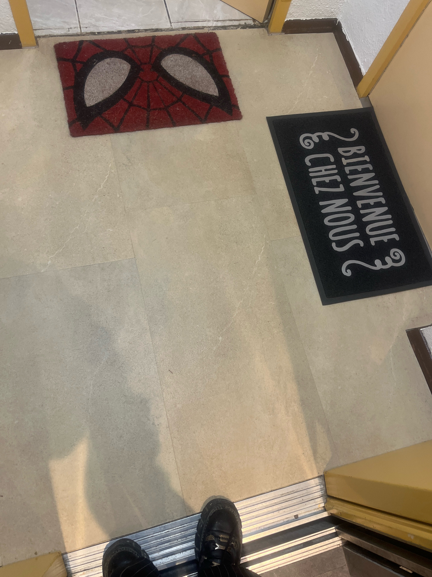 The image shows an indoor scene with two doormats on a tiled floor. The first doormat, positioned on the left, features a red and black design resembling the mask of the superhero Spider-Man. The second doormat, on the right, is black with white lettering that reads "BIENVENUE CHEZ NOUS," a French phrase meaning "Welcome Home." The floor appears to be made of large, light-colored tiles with faint veining. In the foreground, two pairs of feet are visible, clad in dark shoes, standing on a metal threshold, suggesting the person or people are either entering or exiting a space. The environment appears to be an entryway or hallway, with a yellow doorframe visible on the right side of the image. The lighting suggests an indoor setting with some ambient light.