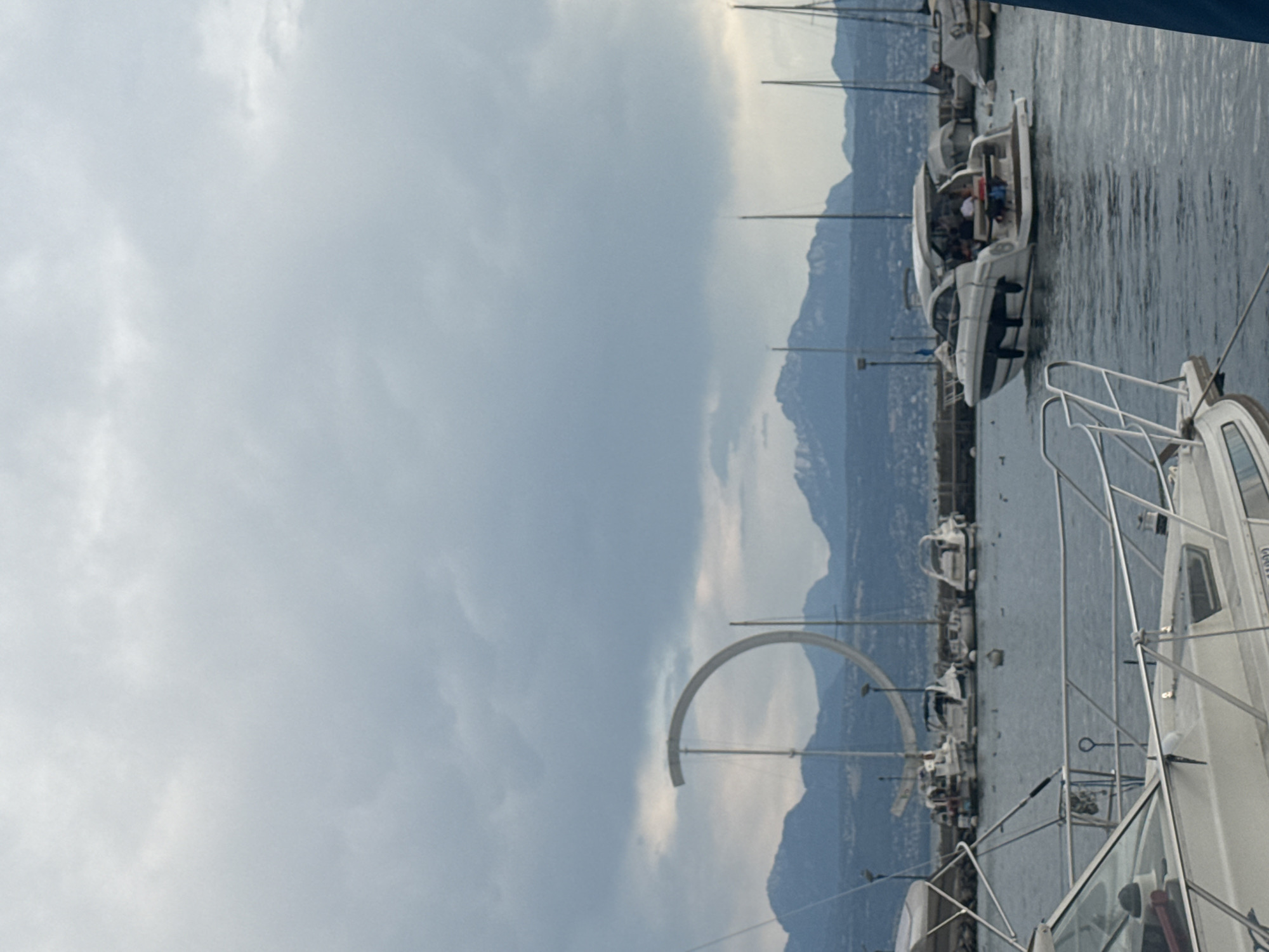 The image shows a harbor with several boats docked and one boat moving. In the background, there are mountains and a large C-shaped sculpture. This sculpture is the Jet d'Eau, a famous landmark in Lausanne, Switzerland. The sky is overcast, and the water appears choppy. The boat in the foreground is a white motor yacht, and another similar yacht is moving in the middle ground. Several other smaller boats and sailboats are visible in the background. The scene appears to be a public recreational area.