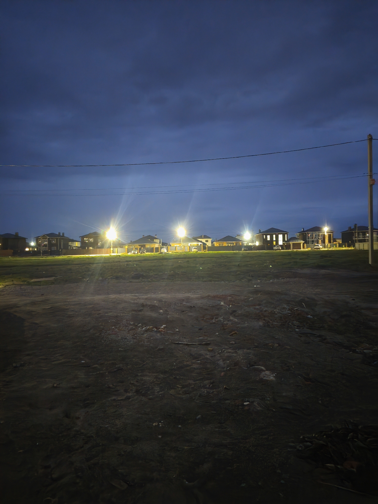 The image depicts a night scene in Сокуры, Russia. In the background, a row of houses is illuminated by streetlights, with visible vehicles in the driveways. An electrical pole with visible wiring is in the top right. The foreground is a dark, unpaved area with scattered debris. The sky is dark, with some cloud cover. No people are visible in the image.