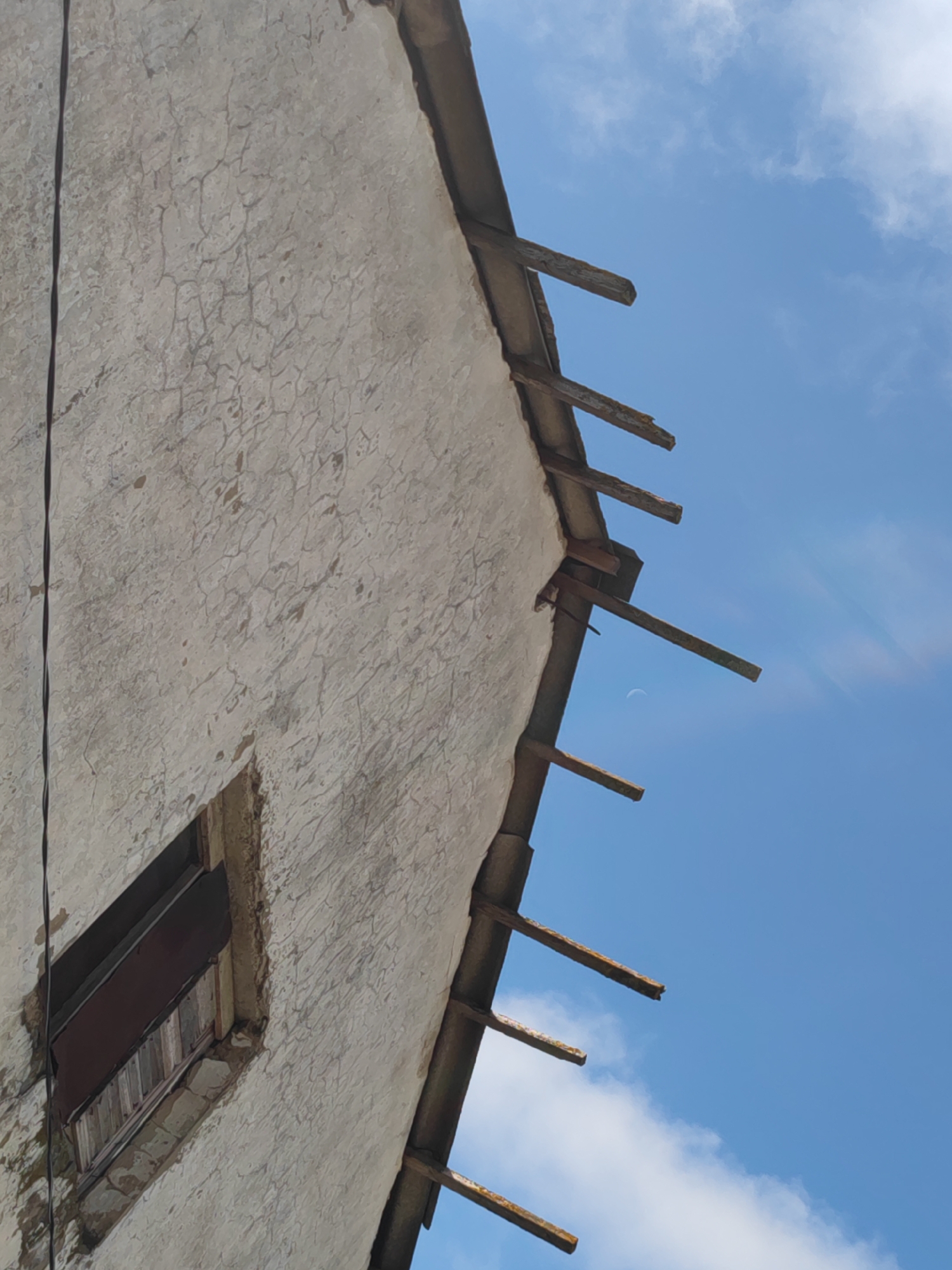 The image depicts the exterior of a dilapidated building in Sennoy, Russia. The building's facade is heavily cracked and weathered, with a single visible window boarded up. The roofline is in disrepair, with exposed and decaying wooden rafters. The background reveals a clear blue sky with scattered white clouds. A power line runs vertically along the left side of the image.