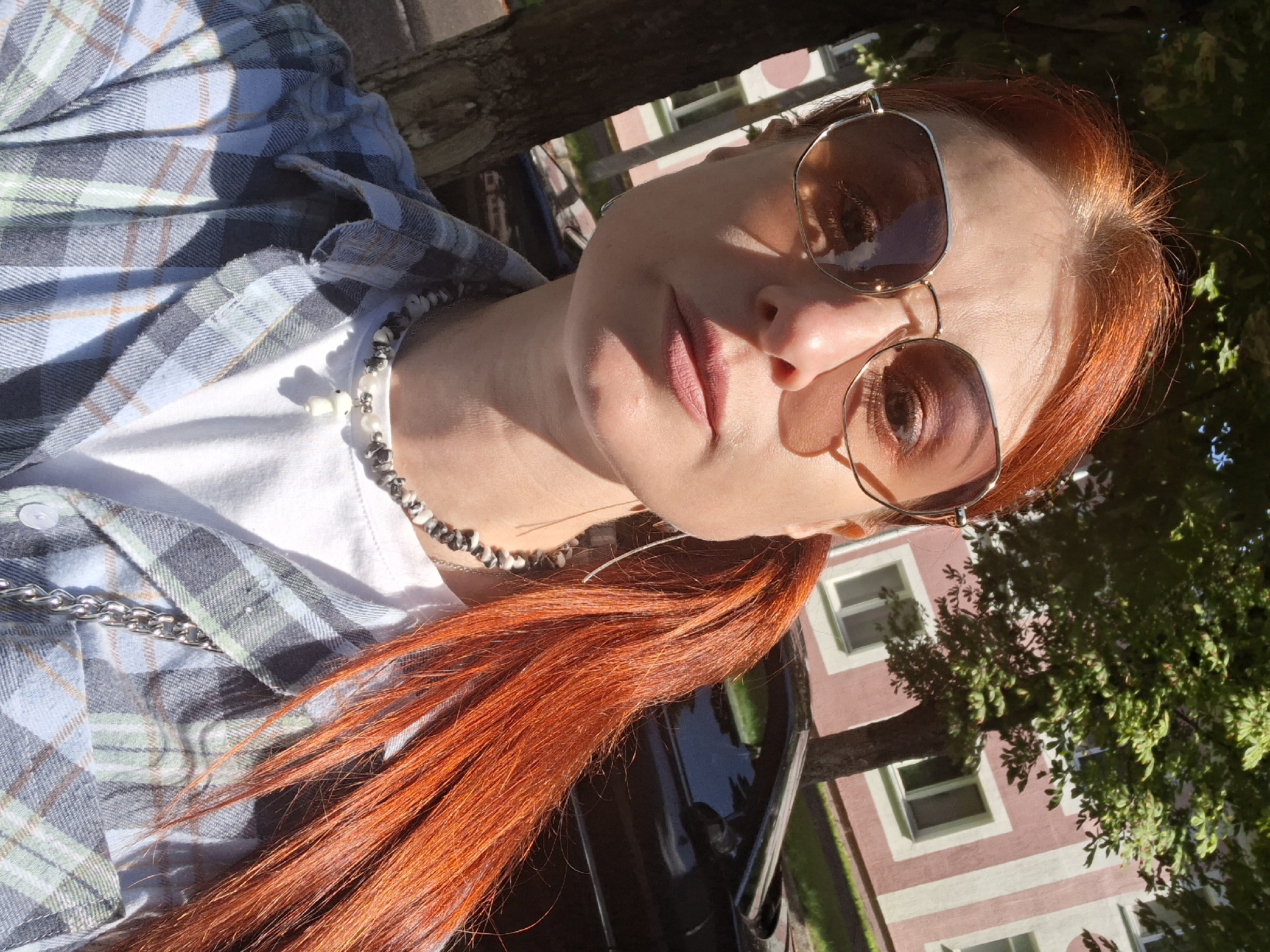 A fair-skinned woman with bright red hair styled in a ponytail and wearing sunglasses is taking a selfie outdoors. She is dressed in a light blue and grey plaid button-up shirt over a white t-shirt and a beaded necklace with a white charm. Behind her, a black car is parked next to a light pink and white building, with a tree in the foreground. The scene is lit by bright sunlight, casting shadows. This appears to be a residential street in the city of Myadzyel, Belarus, given the architectural style of the building and the presence of trees and a parked vehicle.