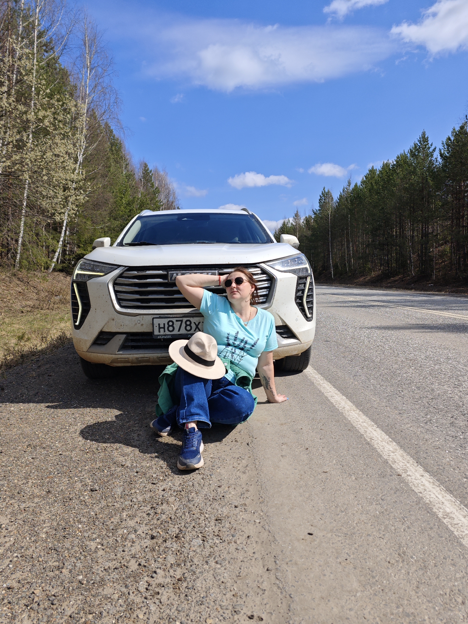 A woman is sitting on the edge of a paved road, leaning against the front of a white SUV with a license plate that reads "H878X". The location appears to be on a rural road outside Chaikovsky, Russia, with trees on either side. The sky is blue with scattered clouds. The woman is wearing a light blue t-shirt, dark blue jeans, and blue sneakers. She is also wearing sunglasses, and a hat is sitting next to her on the ground. A green jacket is draped over her shoulders. She has one arm resting on the car's hood.