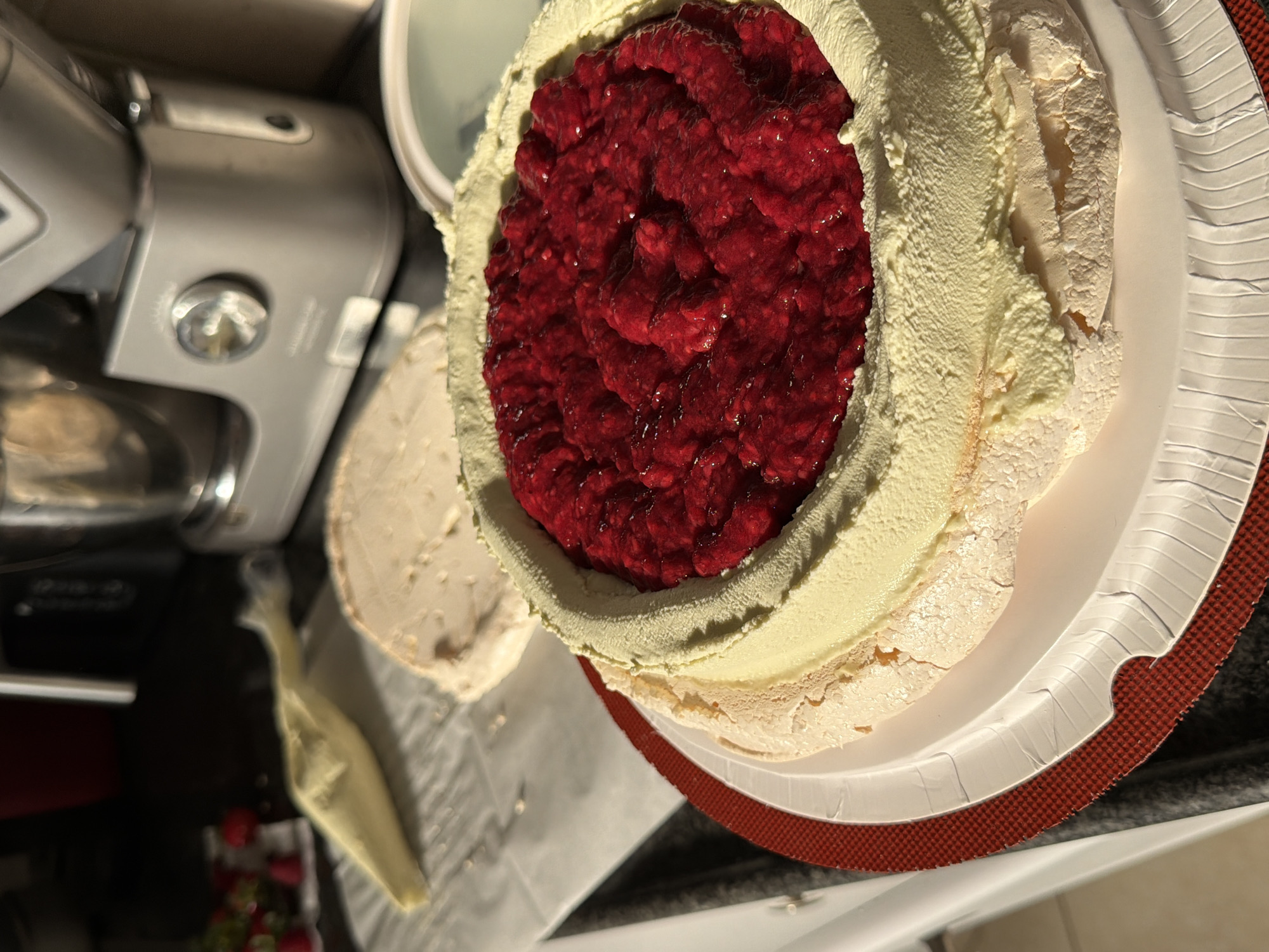 An overhead shot captures a large, round dessert, likely a pavlova or meringue cake, filled with a vibrant red, chunky fruit compote, appearing to be raspberries. The dessert features an outer layer of baked, off-white meringue with visible cracks, and an inner rim of creamy, pale yellow topping, possibly whipped cream or custard, piped around the fruit.
The dessert rests on a white, ridged paper plate, which is placed on a textured red placemat or mat. In the background to the left, a silver or grey stand mixer is visible, showing a control knob and part of its mechanism. Further to the lower left, a small pile of fresh red strawberries is discernible on what appears to be parchment paper, alongside a partially visible piping bag containing a light-colored cream. Another container lid is partially seen in the upper background. The setting suggests an indoor kitchen or dining area, illuminated by artificial light. No clear text is visible in the image.
