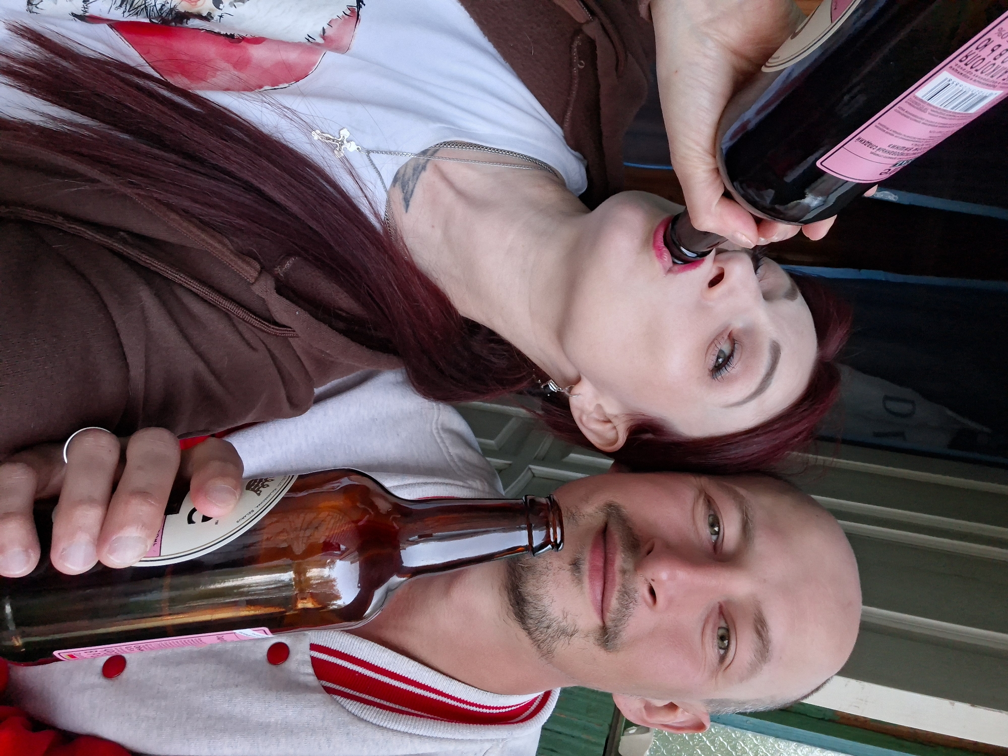 In a close-up shot, a man and a woman are positioned side-by-side, their heads close together. The man, with a shaved head and goatee, holds a brown beer bottle to his mouth. The woman has red hair and is holding a dark wine bottle to her mouth. They are both looking up towards the camera. The location is presumed to be Забалоцкі сельскі Савет, Belarus.