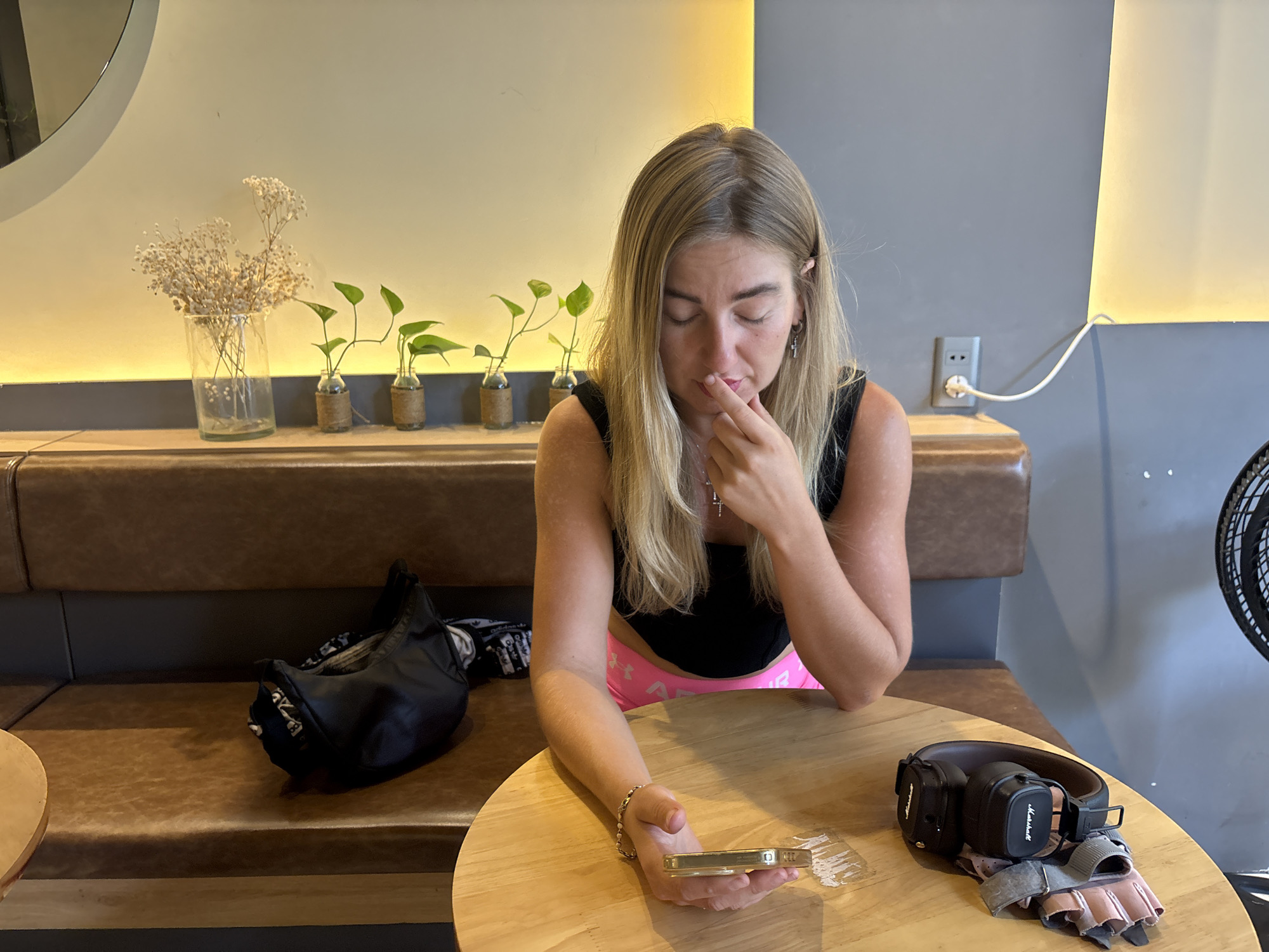 A blonde woman sits at a round wooden table in a cafe. She wears a black tank top, and pink shorts are visible. She holds a gold-colored smartphone in her left hand and touches her lower lip with her right forefinger, gazing downward. On the table are a pair of black Marshall headphones and gray workout gloves. Behind the woman is a brown bench seat. Along the seatback are several small glass vases containing greenery and a larger vase with dried flowers. A black bag sits on the bench to her left. The walls are two-toned, with a yellow stripe above gray. An electrical outlet is visible on the gray portion of the wall. Part of a black fan is visible on the right side of the image. This cafe is located in Đà Nẵng, Vietnam.