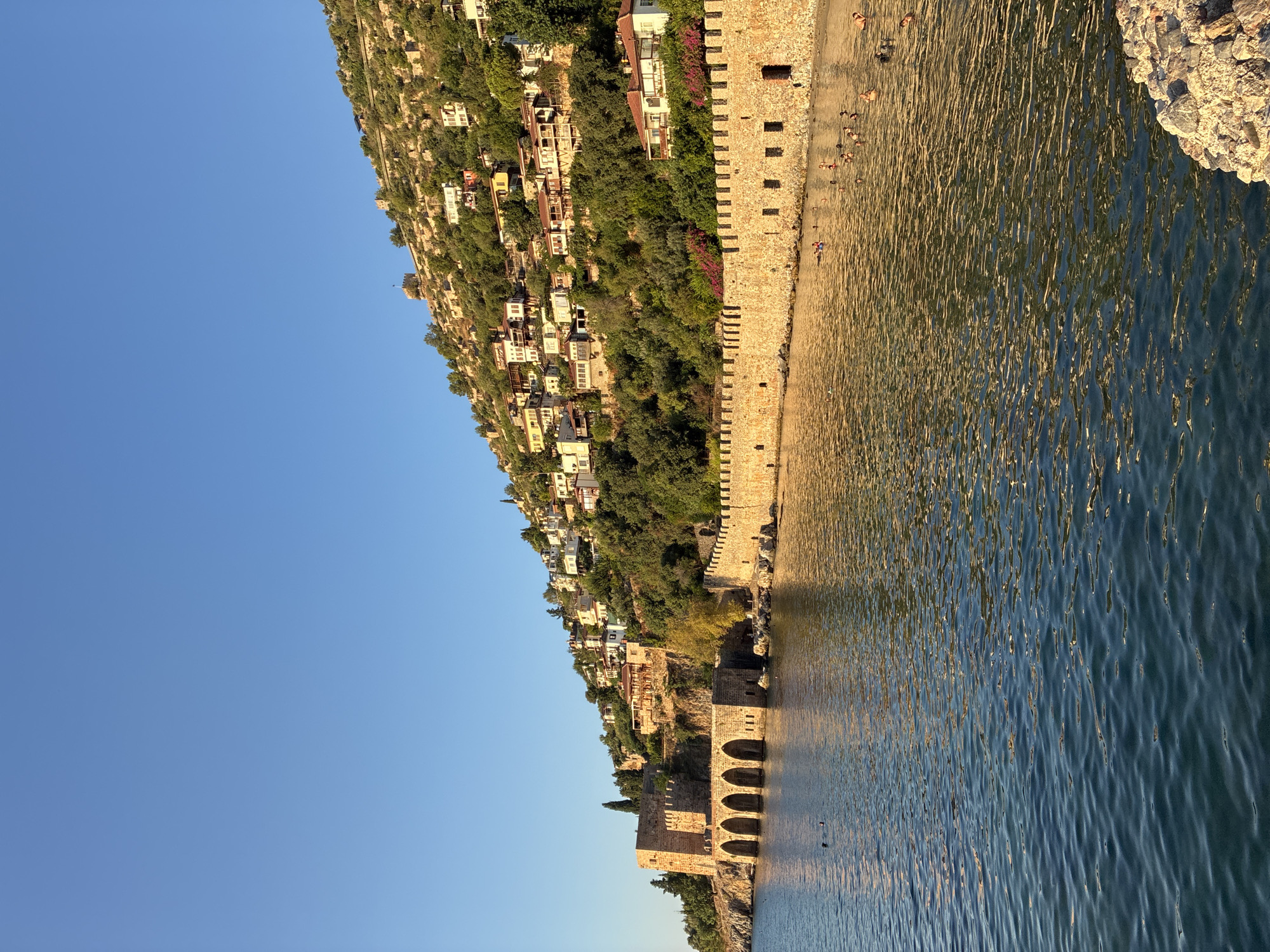 The image depicts the historic Alanya Castle and its surrounding defensive walls, located in Alanya, Turkey. The castle complex is perched on a hill overlooking the Mediterranean Sea. In the foreground, the calm blue water of the sea is visible, with several people swimming and enjoying the water. Along the coastline, a long, stone defensive wall stretches across the frame, part of the castle's fortifications. Behind this wall, the hillside is covered with lush green trees and dotted with traditional white and red-roofed houses, characteristic of Alanya's old town. A prominent stone structure, likely part of the castle's historical architecture, with arched openings, stands near the water's edge on the left side of the image. The sky above is a clear, bright blue, indicating fair weather. The sunlight casts a warm glow on the scene, highlighting the textures of the stone walls and the rippling surface of the water.
